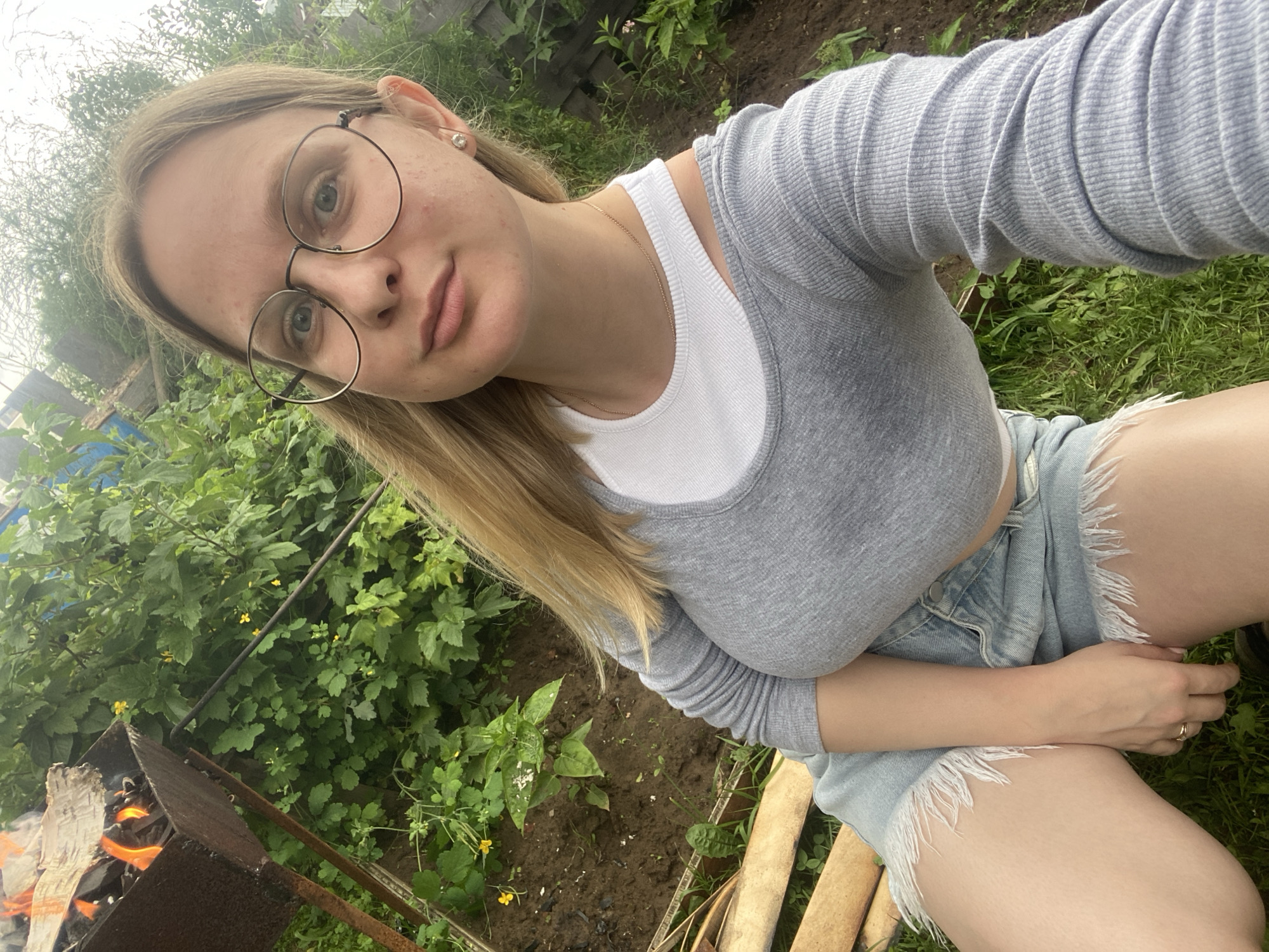 The image depicts a young woman with blonde hair and glasses sitting outdoors in a garden. She is wearing a grey long-sleeved shirt over a white tank top and denim shorts. The woman is holding her phone and taking a selfie, with her face tilted towards the camera. She has a neutral expression on her face.
The background shows lush green foliage and some visible tree branches. There is also a fire pit with burning wood and some gardening tools. The ground is covered with dirt and grass. The lighting suggests it is daytime, possibly overcast, given the diffused light. The overall setting appears to be a casual outdoor space, possibly a backyard or a rural garden area. The presence of the fire pit and gardening tools suggests some outdoor activity is either taking place or has recently occurred. The woman's attire and the setting imply a relaxed, informal atmosphere.