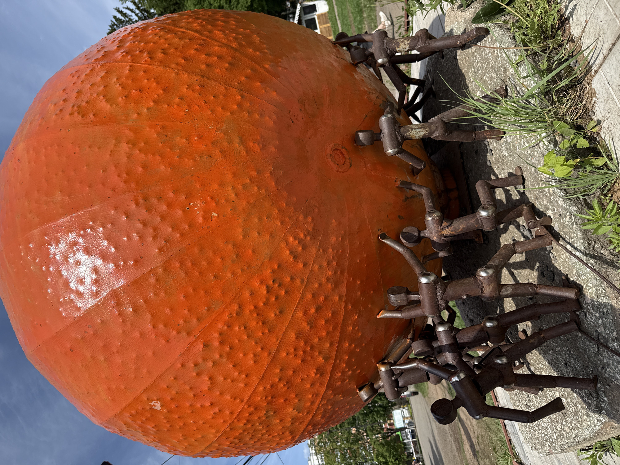 The image shows a large, orange, spherical sculpture, resembling a pumpkin, resting on a base composed of metal figures arranged around its circumference. The sculpture has a textured surface. The base sits on a concrete surface, with grass and vegetation growing nearby. In the background, there are glimpses of buildings, a street, and trees, suggesting an urban or suburban environment. The photograph seems to be taken outdoors during daylight hours. While this particular sculpture is known to be located at the Izhevsk Circus in Izhevsk, Russia, this information cannot be definitively extracted from the image itself.