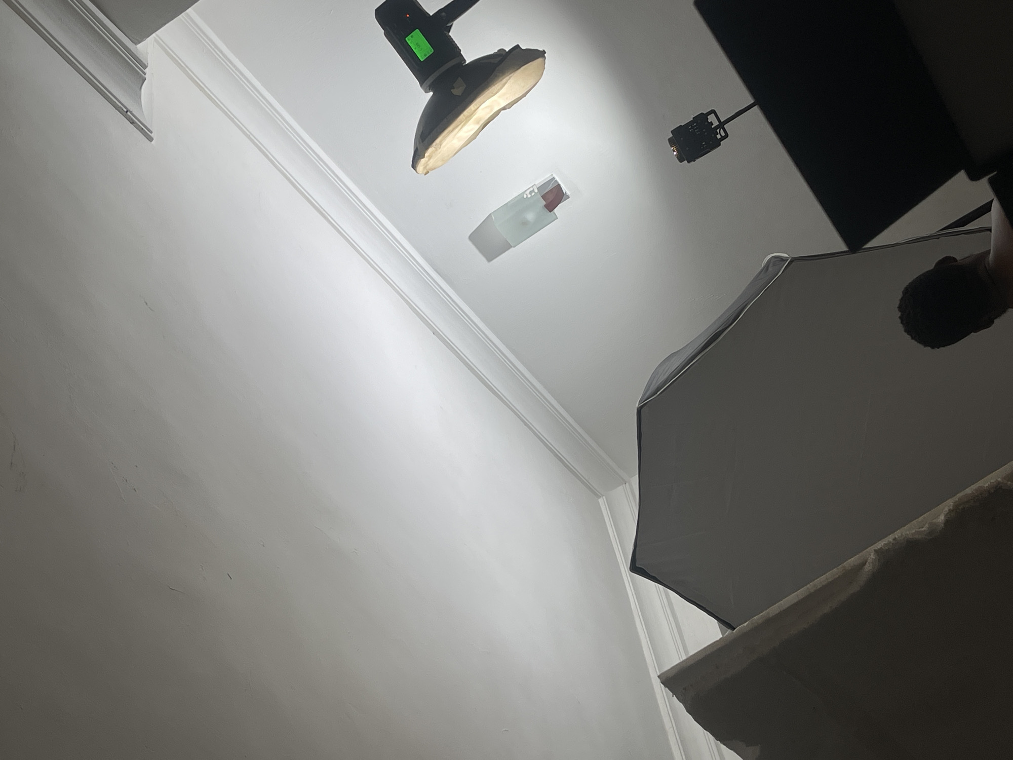 This image captures an indoor product photography setup, seen from a low angle looking upwards towards the ceiling. The scene features multiple lighting elements and a central product.
The primary subject is a translucent, rectangular object, likely a lipstick or cosmetic product, suspended or placed against the white ceiling. It is brightly illuminated by a prominent black spotlight, which is positioned directly above it and casts a strong, focused beam of light. The spotlight has a green digital display on its top, partially visible, showing "8.0" and what appears to be a battery or signal icon.
To the right, a large, dark-grey softbox stands, likely used to provide diffused light. Above the softbox, another smaller, black light fixture is visible. The background consists of clean, white walls and ceiling, adorned with decorative white crown molding along the edges, suggesting a permanent architectural structure.
In the far right of the frame, the top and back of a person's head with dark, curly hair is partially visible, indicating their presence as a photographer or assistant overseeing the shoot. Below the softbox, a light-colored, rough-textured surface can be seen, possibly part of a table or platform. The overall impression is a controlled studio environment designed for precise product lighting and capture. The location context is Dutsen Alhaji, Nigeria.