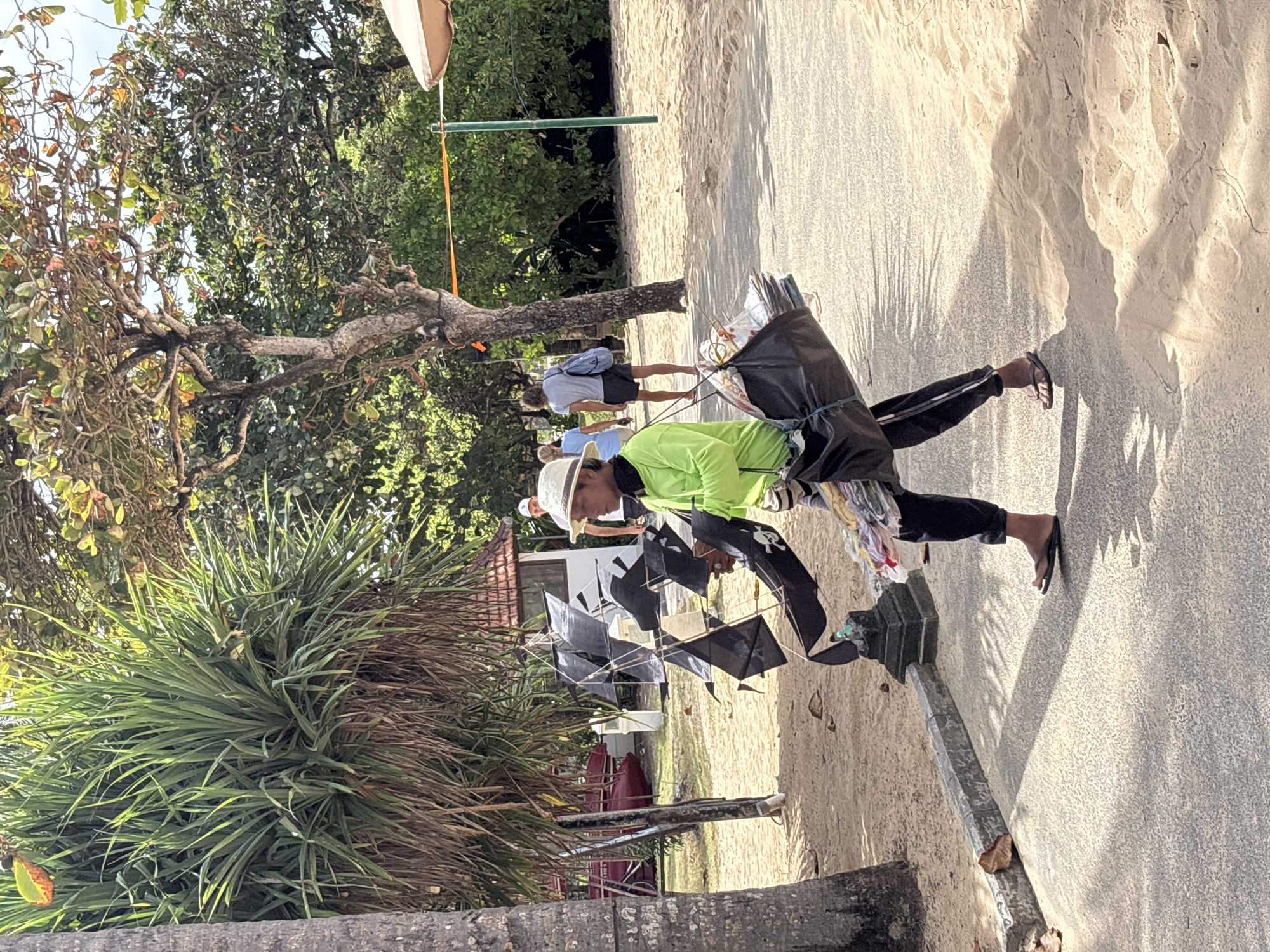 In Kuta Selatan, Indonesia, on a sandy beach path bordered by palm trees and lush foliage, a person wearing a bright green long-sleeved shirt and a straw hat walks from right to left, carrying a collection of black kites on a frame. The kites are stacked and appear to be arranged for sale or display. The person is also carrying a black bag slung over their shoulder, which appears to contain additional items, possibly more merchandise. The ground is a mix of paved path and sand. In the background, other people are visible walking on the beach and there are trees and a small building. The scene suggests a tourist area, likely a beach resort.