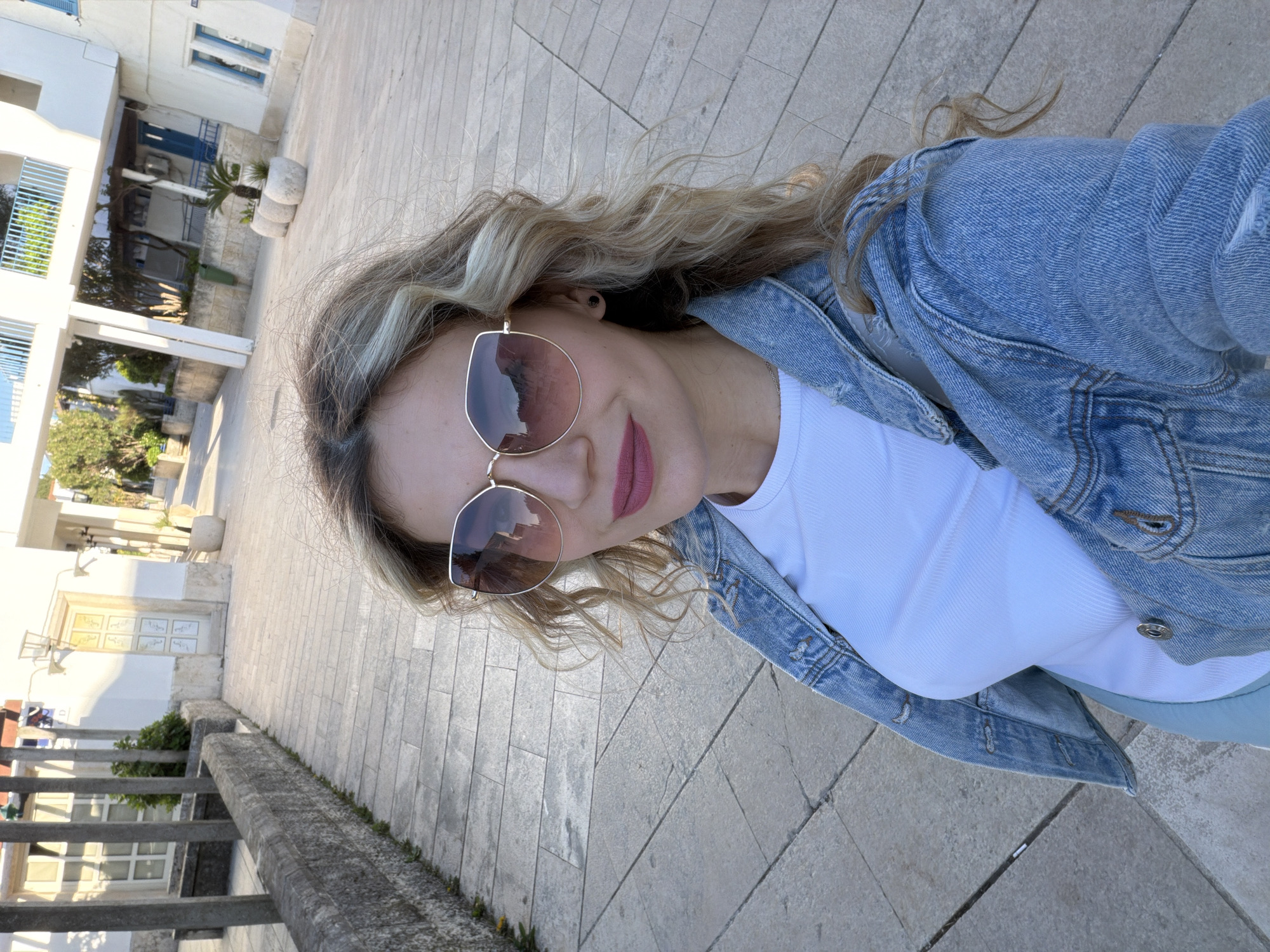 A woman is taking a selfie outside, likely in Budva, Montenegro, given the architectural style. She is wearing sunglasses, a white shirt, and a denim jacket. The background shows a pedestrian area with light-colored stone paving. Buildings with light walls, blue window frames, and railings are visible in the background, typical of Mediterranean architecture.