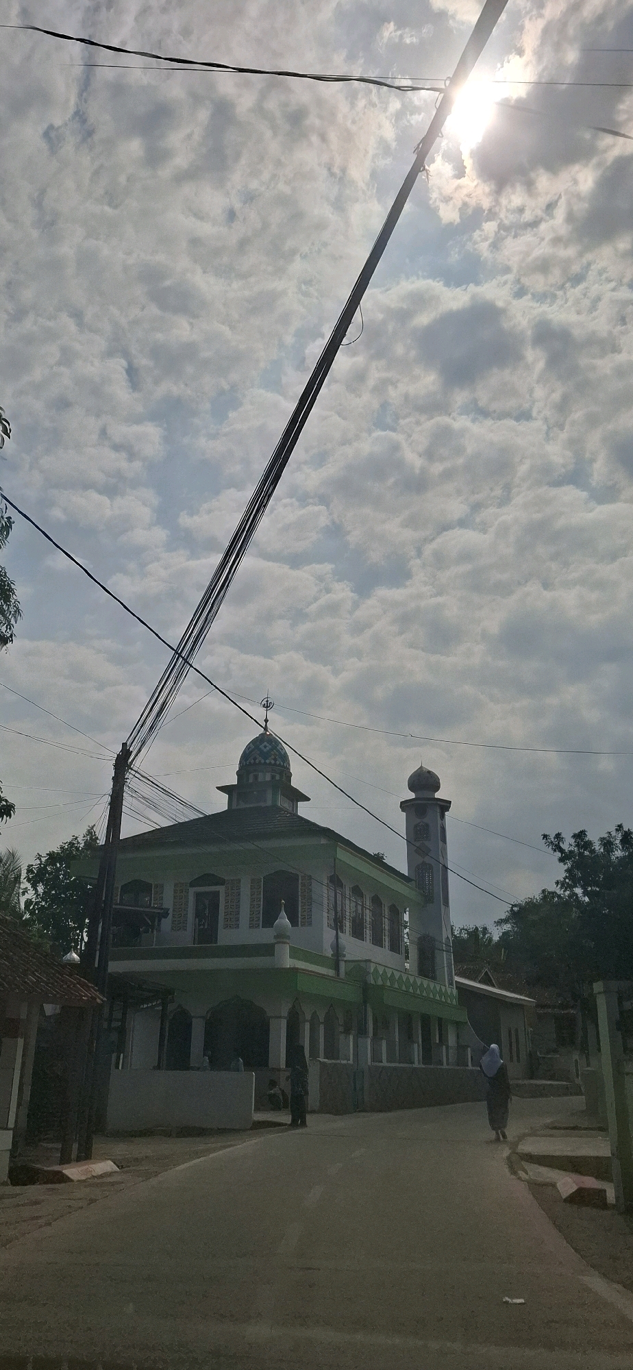 The image captures a mosque in Ciharashas, Indonesia, under a cloudy sky with the sun partially obscured by clouds and a utility pole. The mosque is predominantly green and white, featuring a dome and a minaret. A road runs in front of the mosque, with two individuals visible. One person, dressed in dark clothing, stands near the mosque's entrance, while another, wearing a hijab, walks away from the camera along the road. Several power lines crisscross the sky, with one running diagonally across the frame. The surrounding environment includes trees and other buildings.