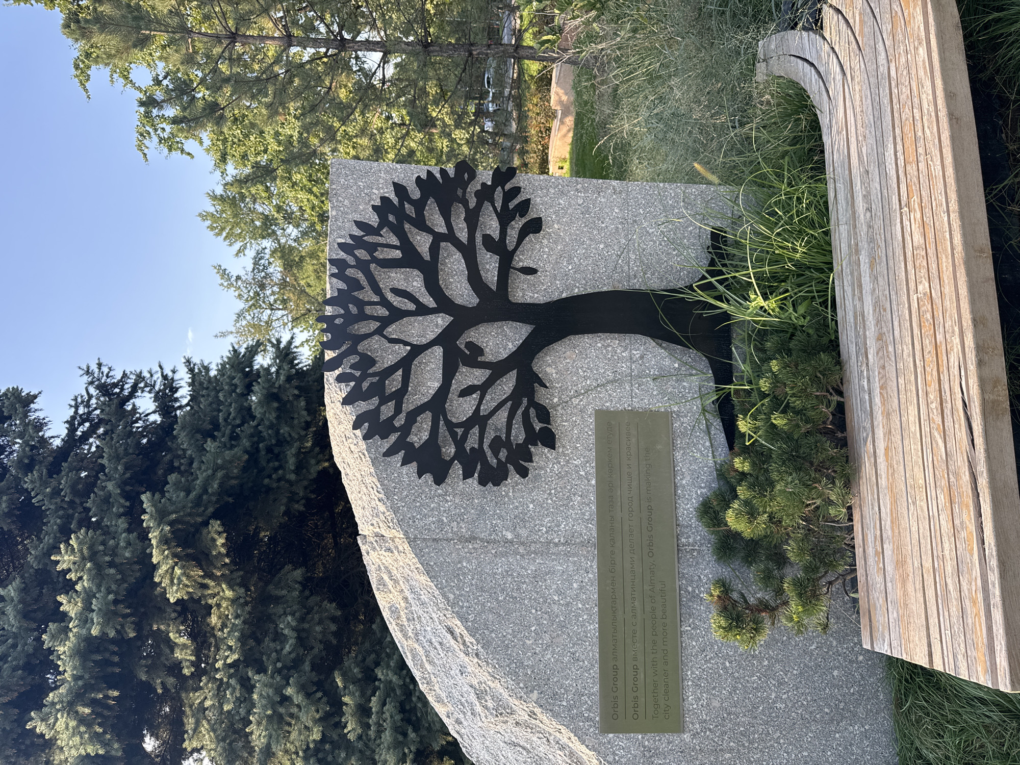 The image shows a monument in a park in Almaty, Kazakhstan. The monument is a large, rough-hewn granite rock with a black metal silhouette of a tree attached to its surface. A plaque is affixed to the rock below the tree, with text in Kazakh, Russian, and English. The English text reads: "Together with the people of Almaty, Orbis Group is making the city cleaner and more beautiful." In front of the monument is a wooden bench, partially curved, with sparse greenery and ornamental grasses around it. In the background, tall evergreen trees and deciduous trees are visible, suggesting a park or natural setting within the city. The sky is clear and blue, indicating a sunny day.