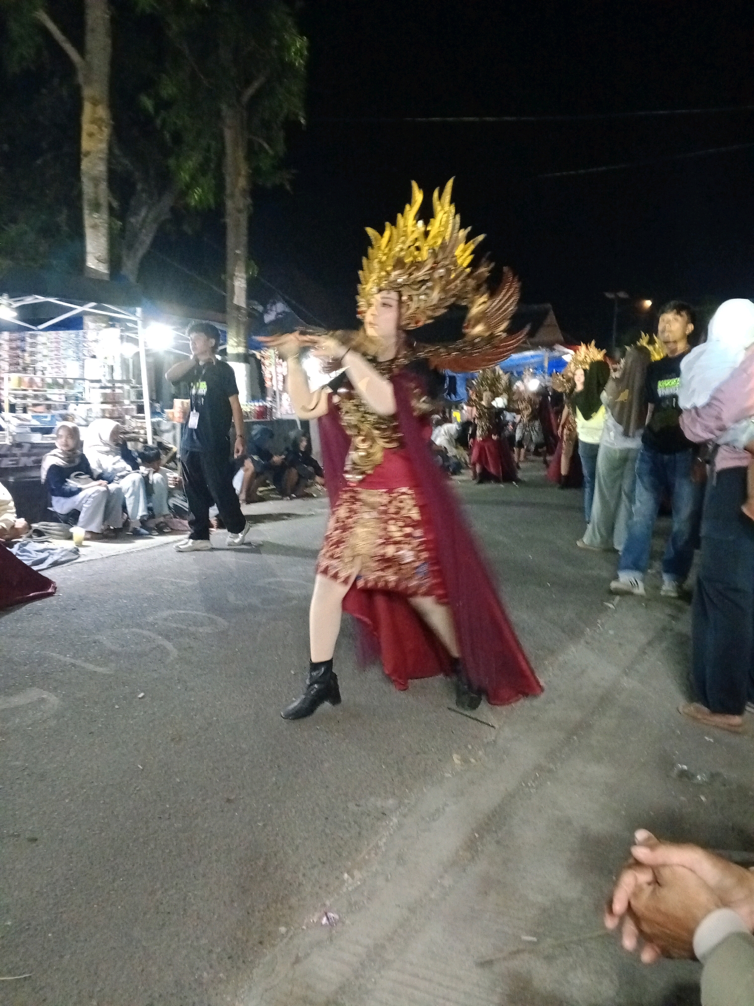 In Druju, Indonesia, at night, a person is performing in a colorful costume, possibly a traditional dancer, wearing a large golden headdress that resembles flames or feathers, a detailed gold and red patterned dress, and a flowing burgundy cape. They are also wearing black ankle boots. The performer is in the middle of the street, with spectators on either side. In the background, several stalls are set up, illuminated by bright lights, and people are gathered around, some sitting on the ground. Trees are visible on the left side of the image, and the sky is dark.