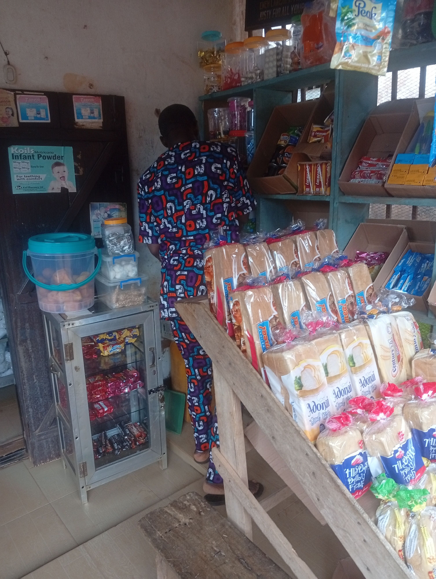 The image depicts a small convenience store, likely in Akokwa, Nigeria, with shelves stocked with various food items and toiletries. A person, wearing a vibrantly patterned shirt and trousers, stands with their back to the camera, facing the shelves.
In the foreground, several loaves of bread are displayed in plastic packaging. Some loaves have white labels with images of bread slices and text that appears to be in a Roman script. Other loaves are wrapped in clear plastic with red and blue accents.
To the left, a silver-colored display cabinet contains more packaged goods, appearing to be snacks or confectionery. Above this cabinet, clear plastic containers hold what seem to be snacks or nuts. A large blue-lidded bucket with a turquoise handle contains more snacks.
The shelves behind the bread display are filled with more packaged goods, including what appears to be canned drinks, small boxes, and bags of items. A prominent bag of "Peak" milk powder is visible on the top shelf. On the wall to the left, several posters or advertisements are displayed, including one for "Koil Matricaria Infant Powder."
The environment appears to be indoors with a simple, somewhat cluttered setup common in small shops. The flooring is tiled. The lighting suggests an indoor setting with natural light coming from the right side. The overall impression is that of a busy, local shop serving everyday needs.