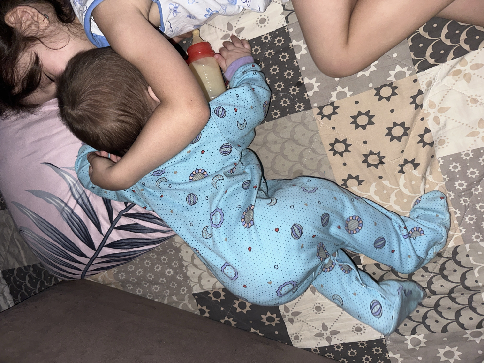A top-down shot shows a child in light blue pajamas printed with moons, hearts, and other small objects lying on a patchwork quilt with a bottle near their head. Another child is partially visible with dark hair, light skin, and wearing a white and blue top. The child in pajamas is nestled against the other child, and a patterned pillow is under their head. The quilt has a variety of light and dark geometric patterns. The location is Unknown, Unknown.