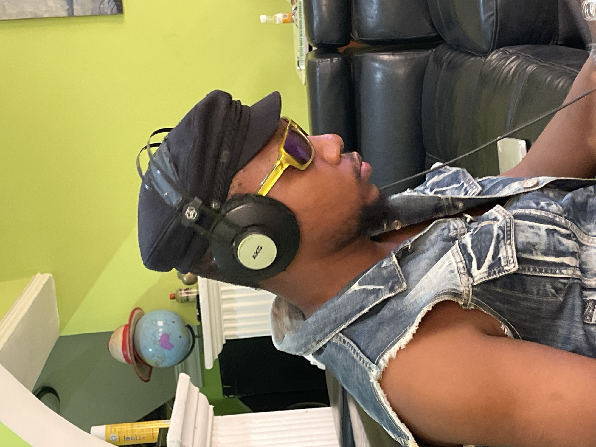 A man wearing a black cap, gold-framed sunglasses, and headphones is wearing a denim vest and is seated in a black leather chair. He is located indoors. The location in Abuja, Nigeria cannot be confirmed from the given image.