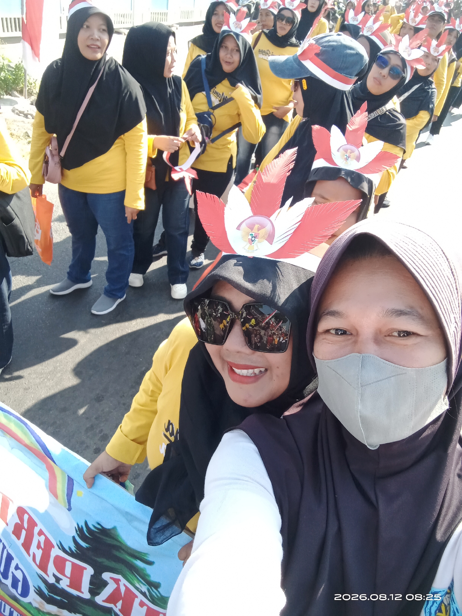In a street scene in Gudo, Indonesia, a group of people, primarily women, are participating in an outdoor event, possibly a parade or celebration. The individuals are dressed in yellow long-sleeved shirts and black headscarves, suggesting a uniform or team attire. Some are wearing festive headwear adorned with red and white feathers and an emblem, likely representing national colors or symbols.
In the foreground, two women are prominently featured. The woman on the left wears sunglasses and a yellow shirt with a black headscarf, her mouth slightly open as if speaking or smiling. She holds a colorful banner with a rainbow motif and text that is partially obscured. To her right, another woman wears a light gray face mask and a dark headscarf, her gaze directed forward.
The background shows more participants in similar attire, some carrying or wearing items related to the event, including Indonesian flags. The environment appears to be an asphalt road with shadows cast by the people and possibly surrounding structures, indicating a sunny day. The overall atmosphere suggests a lively and communal gathering.