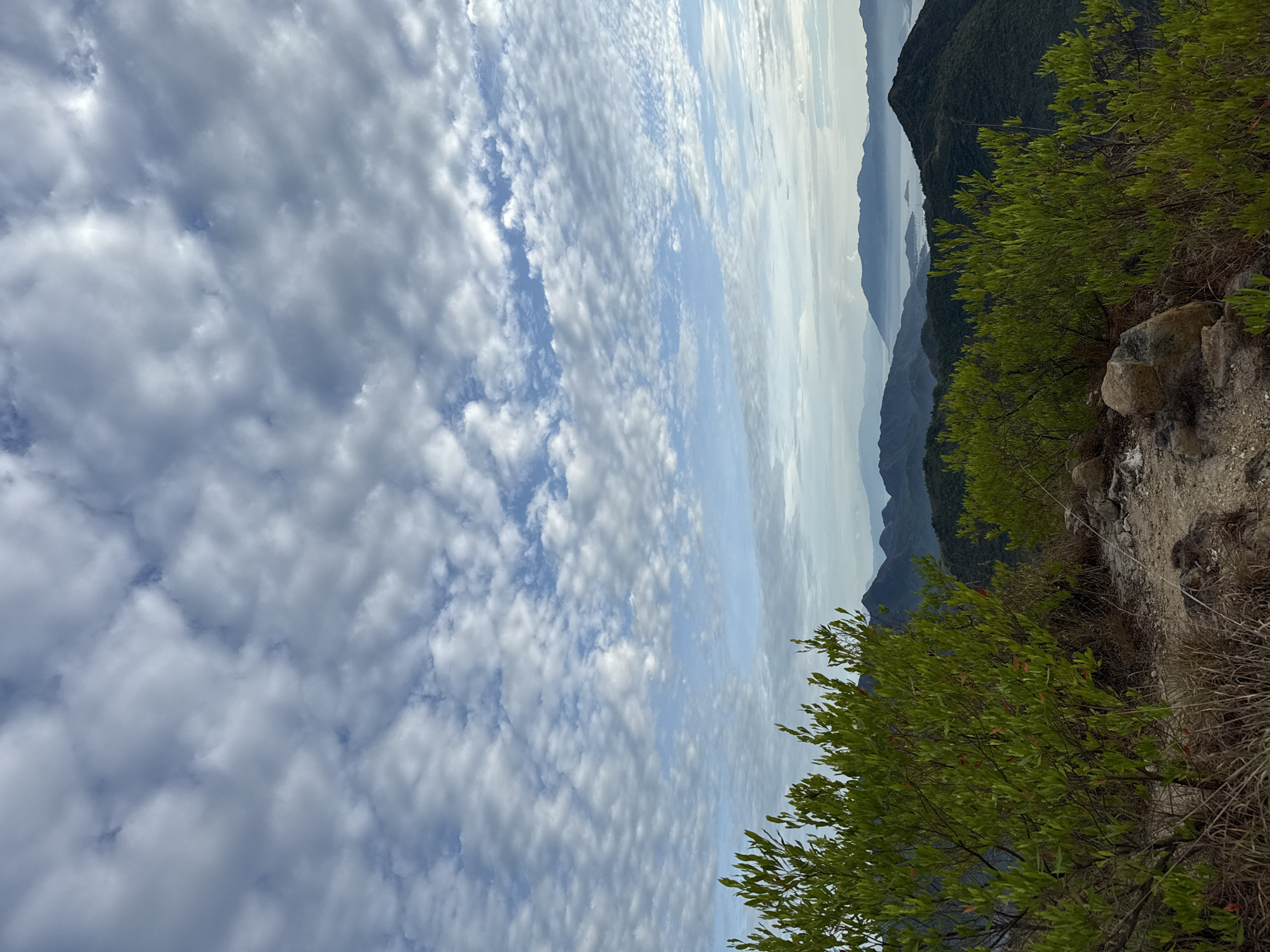 The image depicts a mountainous landscape in Bac Nha Trang Ward, Vietnam. The foreground shows a rocky path with green vegetation and some dried grass. The midground reveals layered mountain ridges in varying shades of gray and green. In the background, the sky is filled with textured clouds, interspersed with patches of blue.
