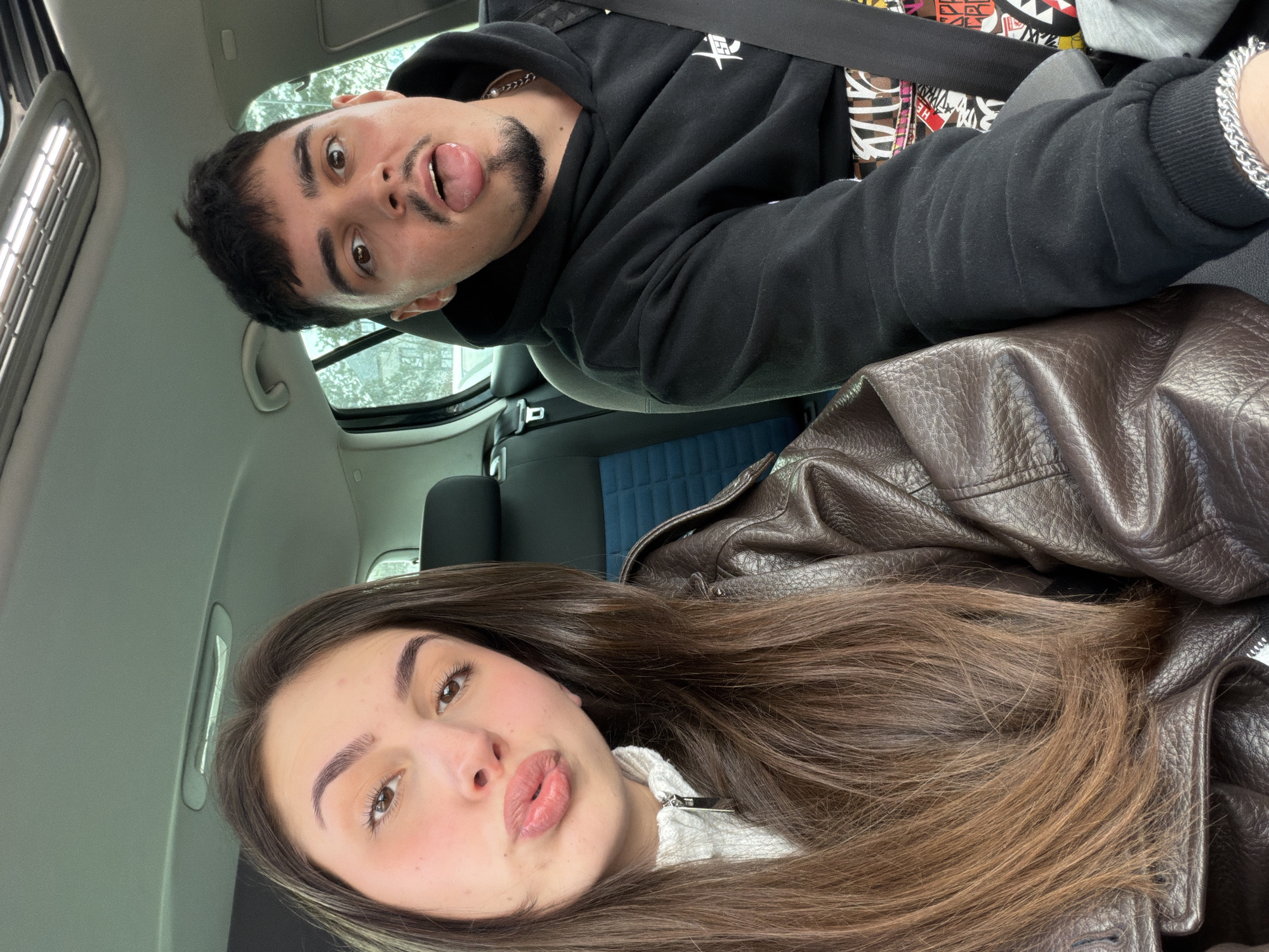 The image captures two individuals inside a vehicle in the city of Sofia, Bulgaria. In the foreground, a female with long brown hair, wearing a white ribbed top with a zipper and a dark brown faux leather jacket, holds the camera for a selfie. Her lips are pursed, and she looks directly towards the camera. To her right and slightly behind, a male with short dark hair, a mustache, and a goatee wears a black hoodie featuring a white stylized Stussy logo on the chest, a silver chain necklace, and is secured by a black seatbelt. He also looks directly towards the camera with his tongue extended. The vehicle's interior includes a visible sunroof mechanism in the ceiling, dark-colored seats, and a blue fabric accent on the side of the front passenger seat. Blurred foliage and structures, indicative of an outdoor urban environment, are visible through the vehicle's windows.