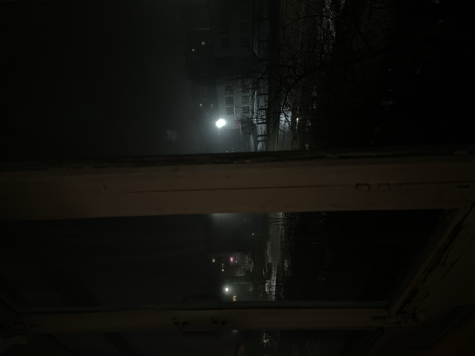 A night scene viewed through a wooden-framed window. Outside, a street is illuminated by streetlights. Bare trees are visible to the right of the window. A building with illuminated windows is in the background. The street appears wet, possibly due to rain or snow. The photograph might have been taken in Chaikovsky, Russia, if provided location is true.