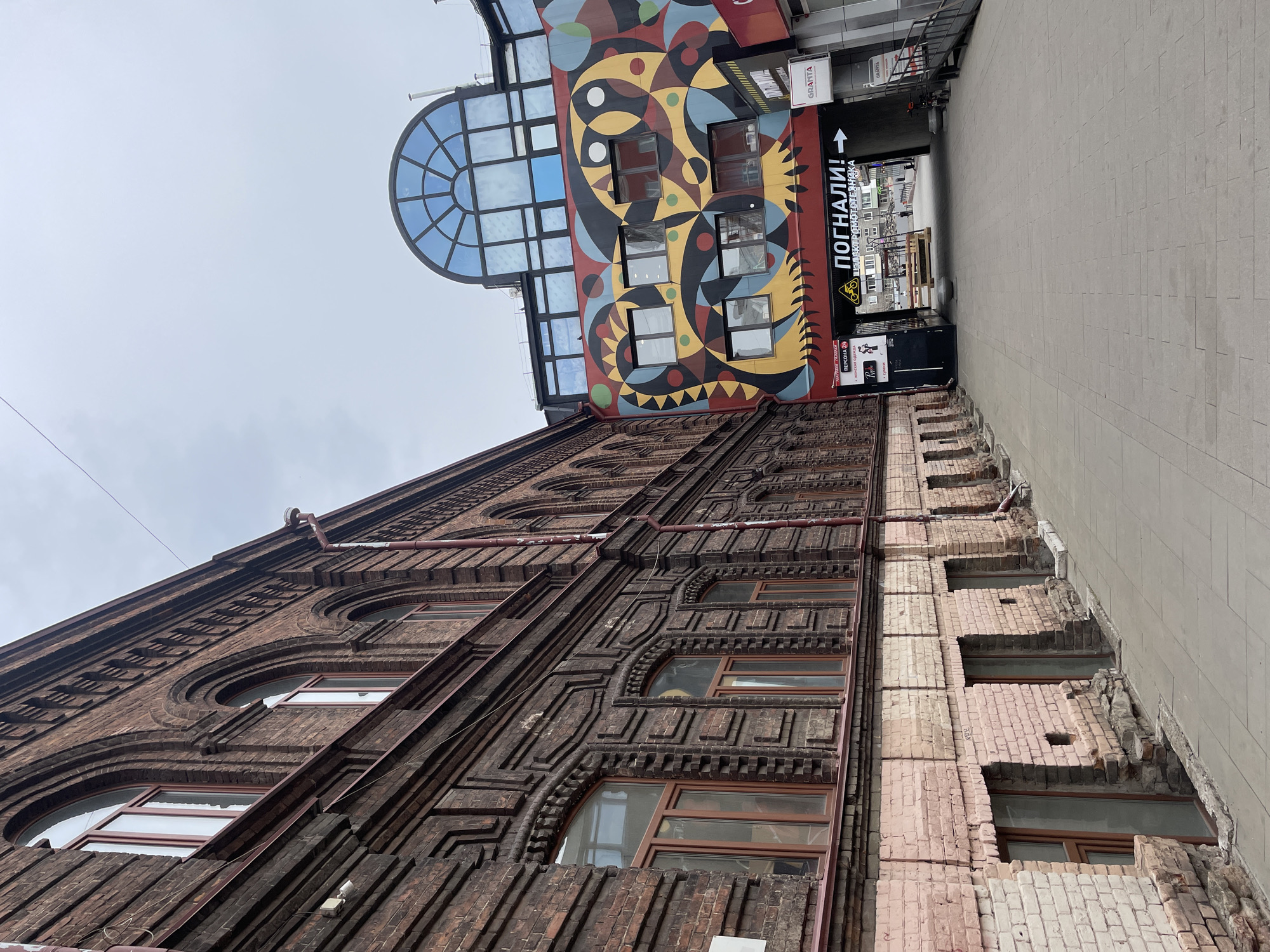 A low-angle photograph captures a narrow urban street or alleyway in Krasnoyarsk, Russia, under an overcast, light grey sky. The scene is dominated by two contrasting building facades.
On the left, an older, multi-story building presents a facade of dark reddish-brown brickwork. It features intricate and elaborate decorative patterns, including arched windows and geometric reliefs around its numerous openings. Some sections of the brick appear lighter, possibly from age or variation in materials.
Further down the street, on the right side, stands a more contemporary building. Its upper section is adorned with a vibrant, abstract mural painted in a striking palette of red, yellow, blue, black, and white. Below the mural, a large, semi-circular arched window, segmented into many glass panes, reflects the cloudy sky. Rectangular windows are also visible below this feature.
At street level on the right, commercial signs are present. A notable sign features an arrow pointing right and the Russian text "ПОГНАЛИ!" (POGNALI! - meaning "Let's go!"). Below it, smaller, less distinct text includes "ТРАДИЦИОННАЯ" (TRADITSIONNAYA - meaning "traditional"). Other visible signs include "GRANTA" and "ПЕРСОНА 24" (PERSONA 24) accompanied by "Женская одежда" (Women's clothing).
The street surface is paved with light grey interlocking pavers, extending into the background where other structures and a gate are faintly discernible. The image does not show any people or specific activities, emphasizing the architectural details and urban environment.