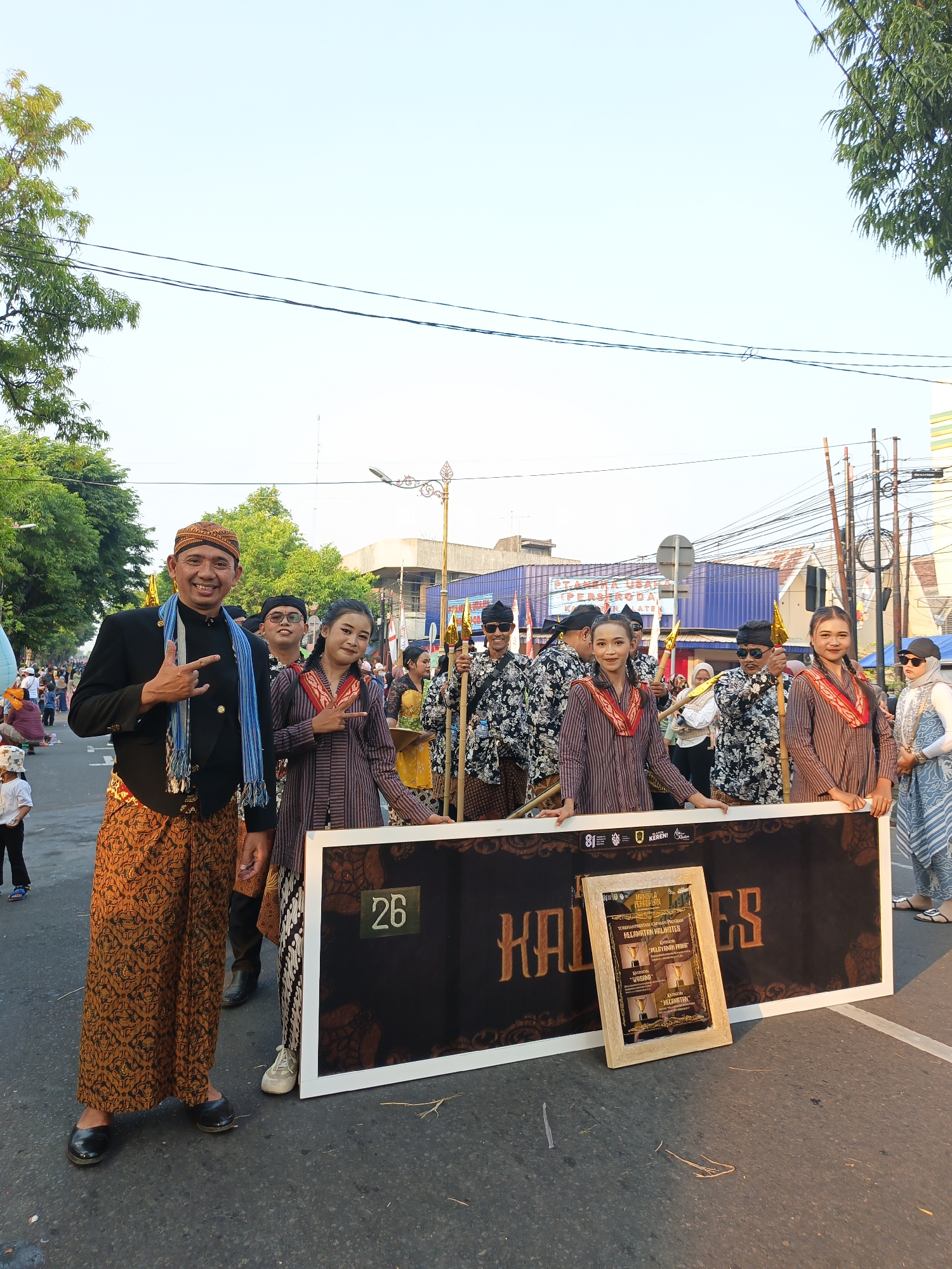 In Klaten, Indonesia, a group of people dressed in traditional Javanese attire pose for a photograph during a street parade. The main subjects are positioned in the foreground, holding a large black banner with "KALILATES" written in decorative, golden letters. A framed certificate or award with Indonesian text is propped up in front of the banner. The people in the foreground are smiling and some are making hand gestures. Behind them, other participants in the parade are visible, also in traditional clothing and carrying spears with golden flags. The environment appears to be a street with trees and buildings in the background, suggesting an outdoor event. The sky is clear and the lighting indicates it is daytime.