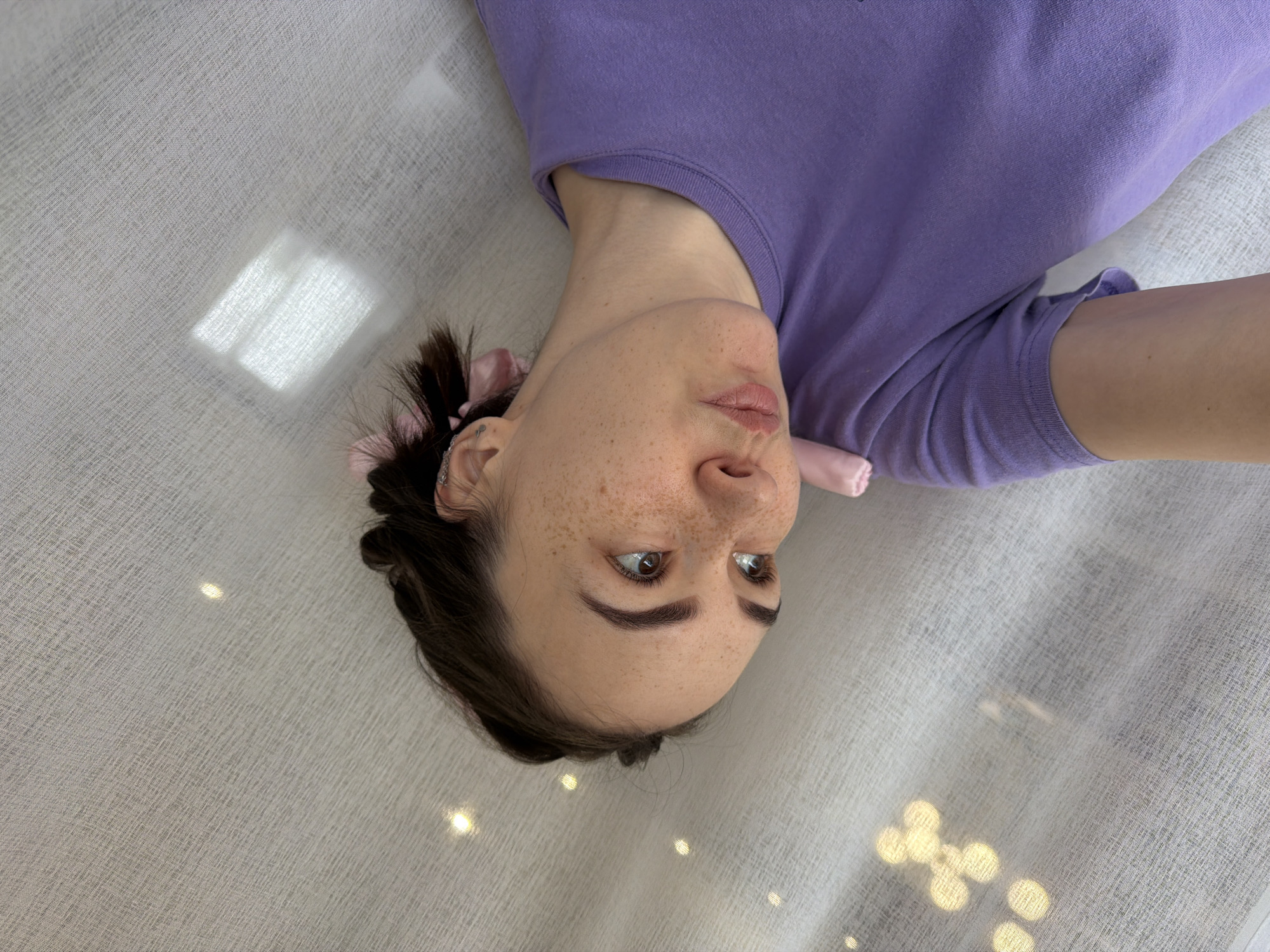 A woman with brown hair and freckles is lying on a light gray fabric surface. She is wearing a purple t-shirt and has pink scrunchies in her hair. The surface she is lying on has some light reflections, possibly from a window. The image was taken from a high angle, looking down at the woman. The location is not discernible from the image.