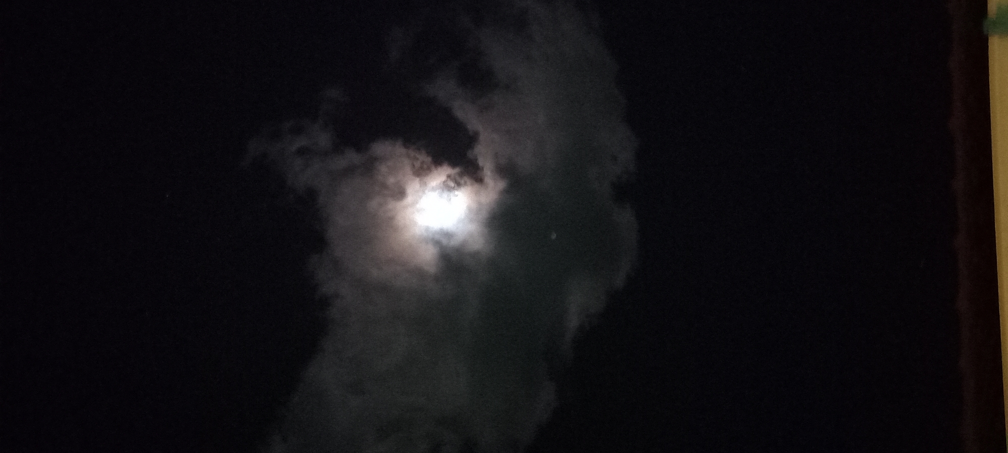 The image presents a nocturnal sky scene from Baan Chaloklum, Thailand, characterized by a bright, moon-like light source partially veiled by clouds. The central feature is a brilliant, diffuse orb of light, likely the moon, which illuminates the surrounding atmospheric conditions. Patches of translucent, wispy clouds, ranging in tone from dark grey to luminous white, are scattered across the frame, with denser formations directly around the light source creating a soft glow. The sky around the clouds and moon is intensely dark, typical of nighttime. On the far right edge of the photograph, a narrow vertical strip of a structure, possibly a wall or window frame, is visible in shades of brown and gold, suggesting the vantage point of the photographer. There are no identifiable people, animals, or specific activities occurring. No discernible text is present. The overall impression is one of a serene, cloudy night sky.