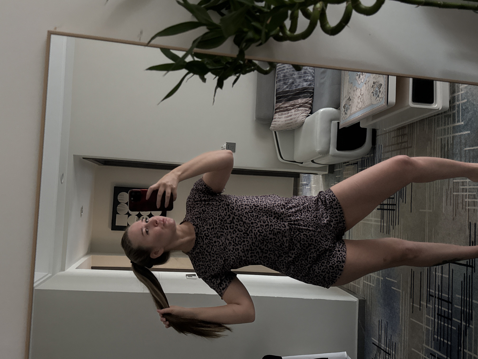 A woman with blonde hair pulled into a high ponytail is taking a mirror selfie in what appears to be a hotel room. She is wearing a leopard-print short-sleeved top and matching shorts. In the reflection, a couch with decorative pillows and a coffee table with a floral print are visible. To the right of the woman, a plant with twisted green stalks is partially seen in the mirror's reflection. The room has a patterned carpet and light-colored walls, with a doorway visible in the background. The overall impression is of a casual moment captured in a domestic setting. The specific location in Yiwu, China, is not determinable from the visual evidence.