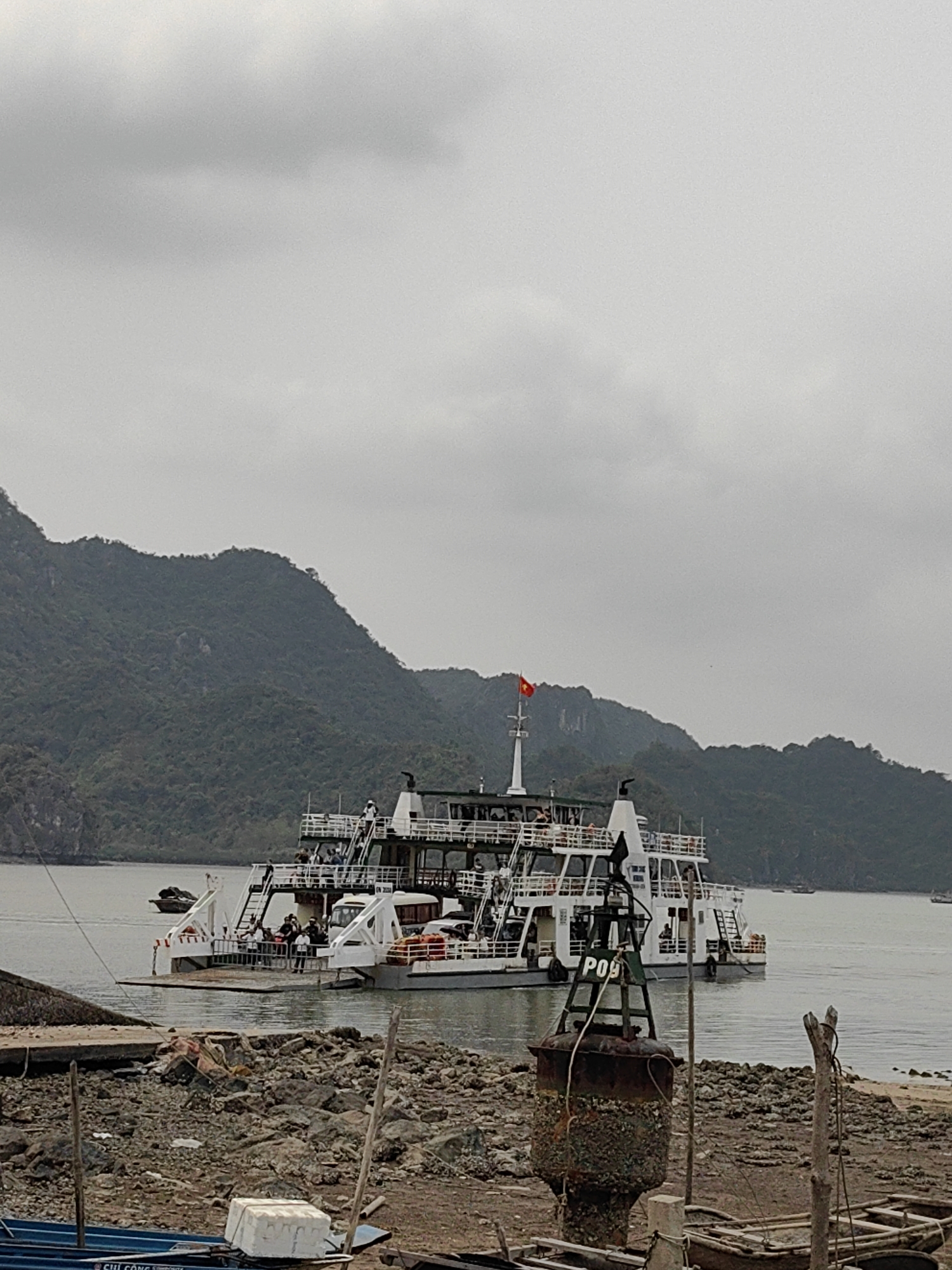 The image shows a multi-deck ferry boat docked near the shore in what appears to be a harbor, likely in Hải Phòng, Vietnam. The boat is white with multiple levels and appears to be carrying passengers. A Vietnamese flag flies on the boat's mast. The surrounding environment includes a rocky shoreline in the foreground. The water is calm, and in the background, there are lush, green, mountainous islands or landmasses. The sky is overcast with heavy grey clouds. A rusty, green buoy marked "P09" sits near the shoreline in the foreground. There are wooden structures and small boats in disrepair closer to the shore as well. The general ambiance of the scene is muted due to the overcast conditions. People are visible on the various decks of the boat. The boat is actively docked with a ramp leading to the pier.