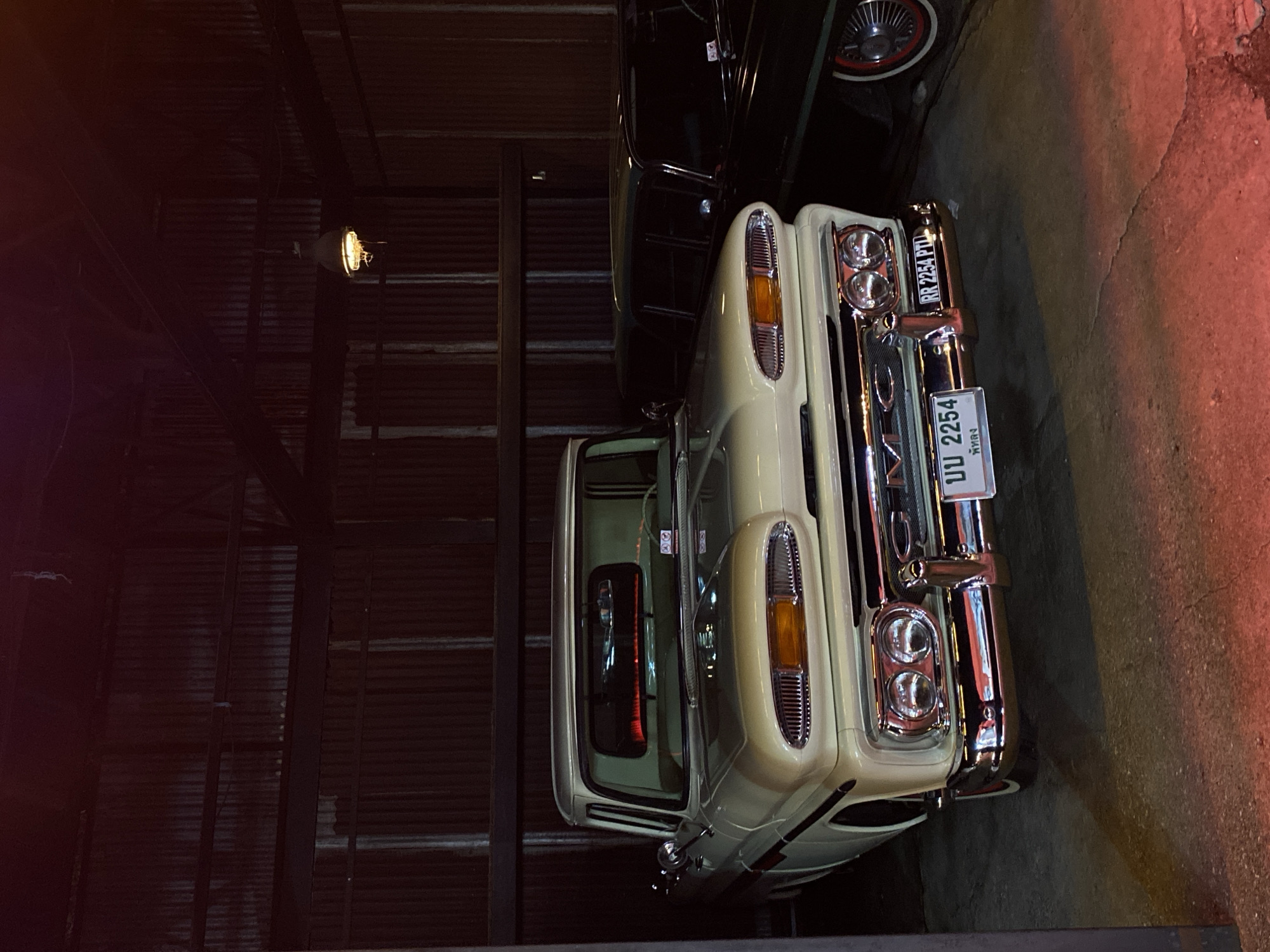 The image shows a close-up view of the front of a vintage, light-colored pickup truck parked indoors. The truck has prominent chrome detailing around its headlights and grille. Its license plate reads "UU 2254," and "Phra Nakhon" is visible beneath the number, suggesting it's registered in the Phra Nakhon district. The location is Bangkok, Thailand. The truck is positioned next to another vintage car, which is dark in color with a visible wheel featuring a white-wall tire. The surrounding environment appears to be a garage or storage space with corrugated metal walls and a single light fixture.
