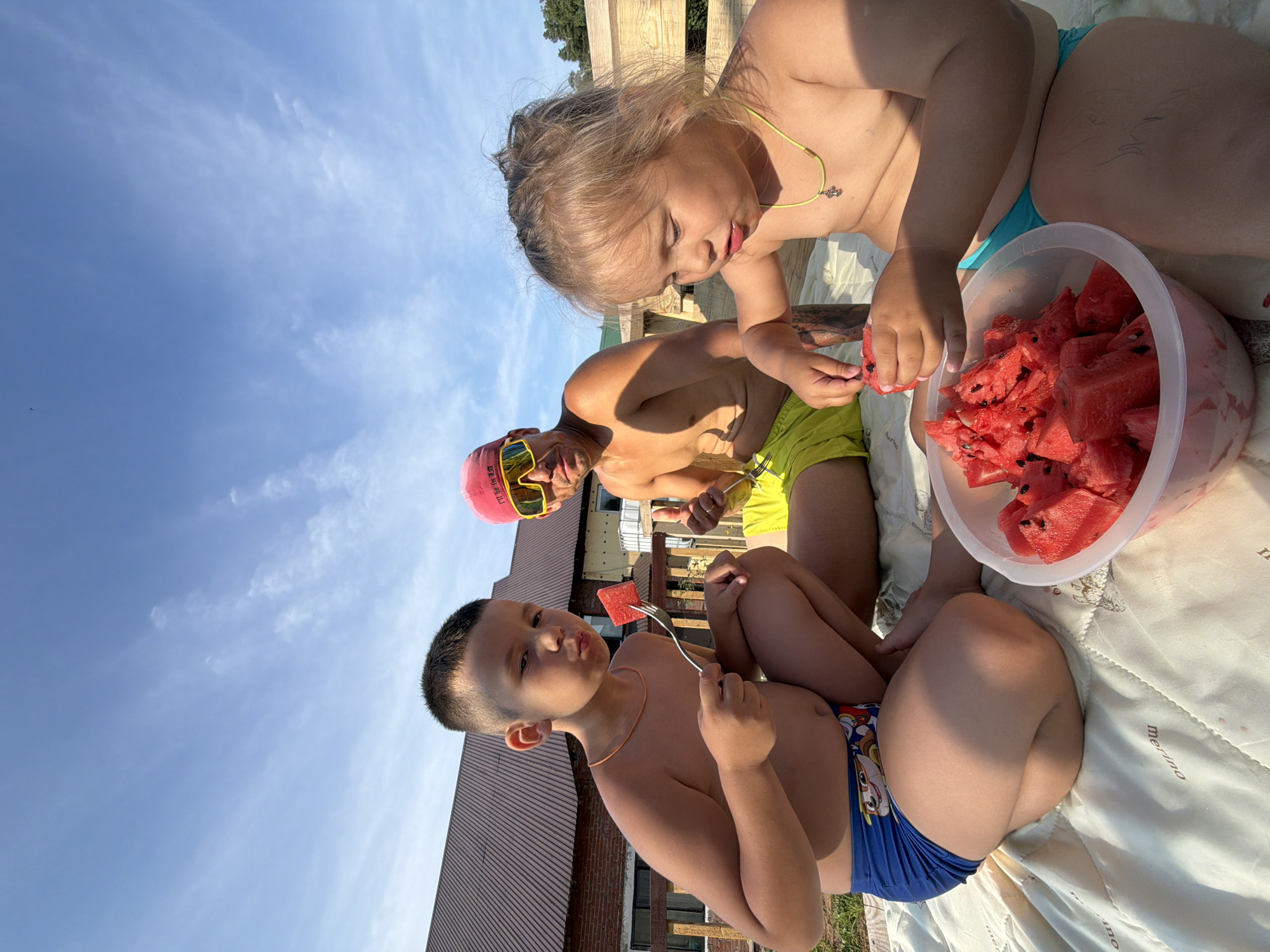 In Chaikovsky, Russia, on a clear day with wispy clouds, three individuals are seated on a blanket outdoors. The scene appears to be in a backyard or a similar residential outdoor space, with a house and fence visible in the background.
On the left, a young boy is kneeling and eating a slice of watermelon from a fork. He is wearing blue swim trunks with cartoon characters. In the center, a man wearing a red cap, yellow sunglasses, and yellow swim trunks is giving a thumbs-up gesture. On the right, a young girl is sitting and eating watermelon, with a necklace around her neck. In front of the children is a large bowl filled with sliced watermelon. The lighting suggests it is daytime, likely afternoon, due to the long shadows cast.