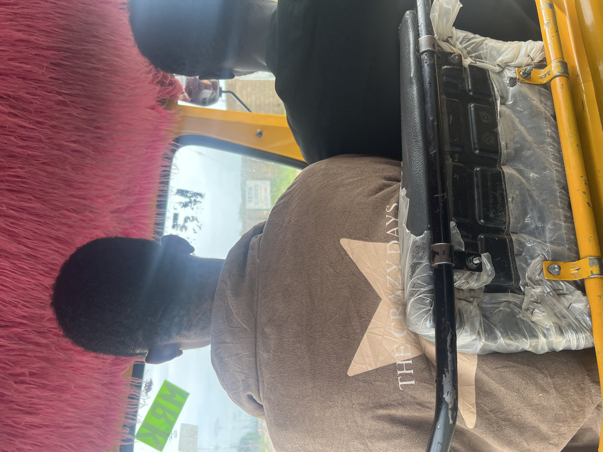In the city of Jalingo, Nigeria, two individuals are seated in the rear of a yellow auto-rickshaw. The interior of the vehicle is decorated with vibrant pink fur lining the ceiling. The person in the foreground is wearing a brown jacket with a large, light brown star graphic and the text "THE CRAZY DAYS" printed on the back. The second individual, to the right, is wearing a black shirt. The auto-rickshaw appears to be in motion, as suggested by the blurred background. The windshield is visible, offering a view of the external environment, which includes some buildings and vegetation. A yellow metal bar is visible across the front of the seating area, covered in clear plastic.