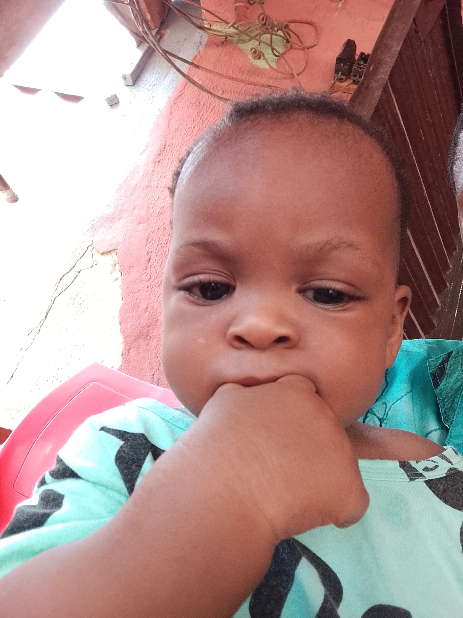 The image presents a close-up portrait of a baby or toddler, likely of African descent, with smooth skin, dark eyes, and short, dark curly hair. The child is wearing a light blue or mint green t-shirt adorned with dark grey or black patterns, possibly depicting stars or abstract shapes. The baby's left hand is raised to their mouth, with fingers or a fist partially inserted, a common gesture for comfort-seeking, teething, or self-soothing. Their expression is neutral, gazing slightly downwards or to the side, conveying a pensive or curious mood.
The child appears to be seated in or leaning against a bright red plastic chair, partially visible on the left side of the frame. A portion of an adult's body, including an arm and shoulder wearing a dark green patterned garment, is visible on the right, suggesting the child is being held or supported. Dark, short curly hair, likely belonging to the adult, is visible in the upper right background.
The background reveals a textured wall with two distinct color sections: a reddish-brown area and a lighter, possibly white or cream-colored section that shows signs of wear and cracking. Exposed electrical wires, some coiled, and small black electrical components or fixtures are visible attached to the wall or a wooden beam in the upper part of the background. Wooden slats or planks are also present on the right side behind the child. The lighting is bright and natural, indicating it is daytime. There is no visible text in the image. The scene suggests an informal setting, possibly outdoors or in a semi-enclosed space.