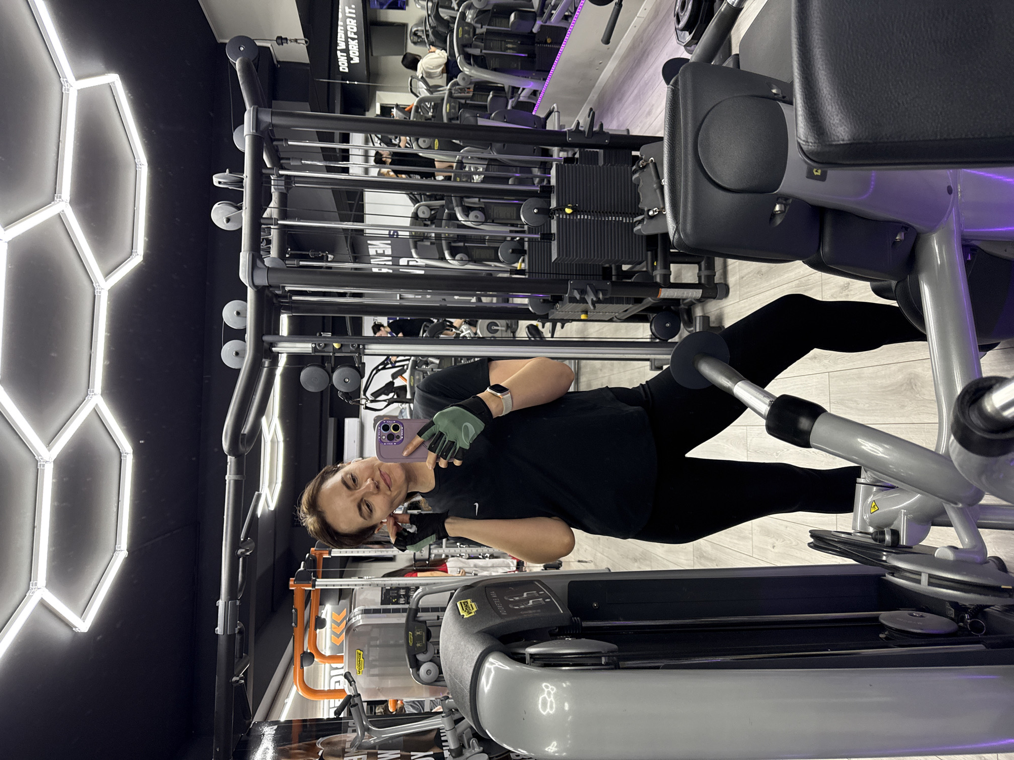 A woman is at a gym in Budva, Montenegro. She stands near a treadmill and weights, holding a cell phone with her right hand, while her left hand makes the peace sign. She wears a black t-shirt and black leggings, a watch, and green and black gloves. The gym has a black ceiling with hexagonal-shaped white lights on the wall.
