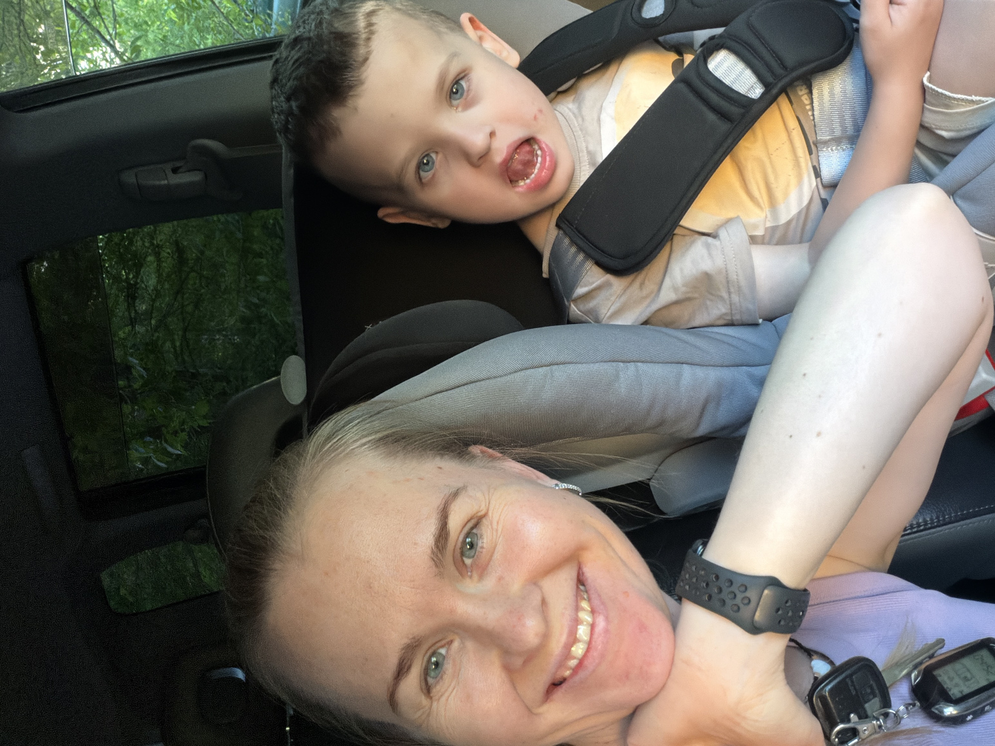 In a car, a woman with blonde hair and blue eyes smiles at the camera, resting her chin on her hand. She is wearing a light purple tank top and a black watch on her left wrist. Next to her, a young boy with short brown hair and blue eyes sits in a car seat, looking forward with his mouth open. He is wearing a tan t-shirt with a yellow design and a car seat harness. The background shows the interior of a car, with trees visible through the windows. The lighting suggests it is daytime.