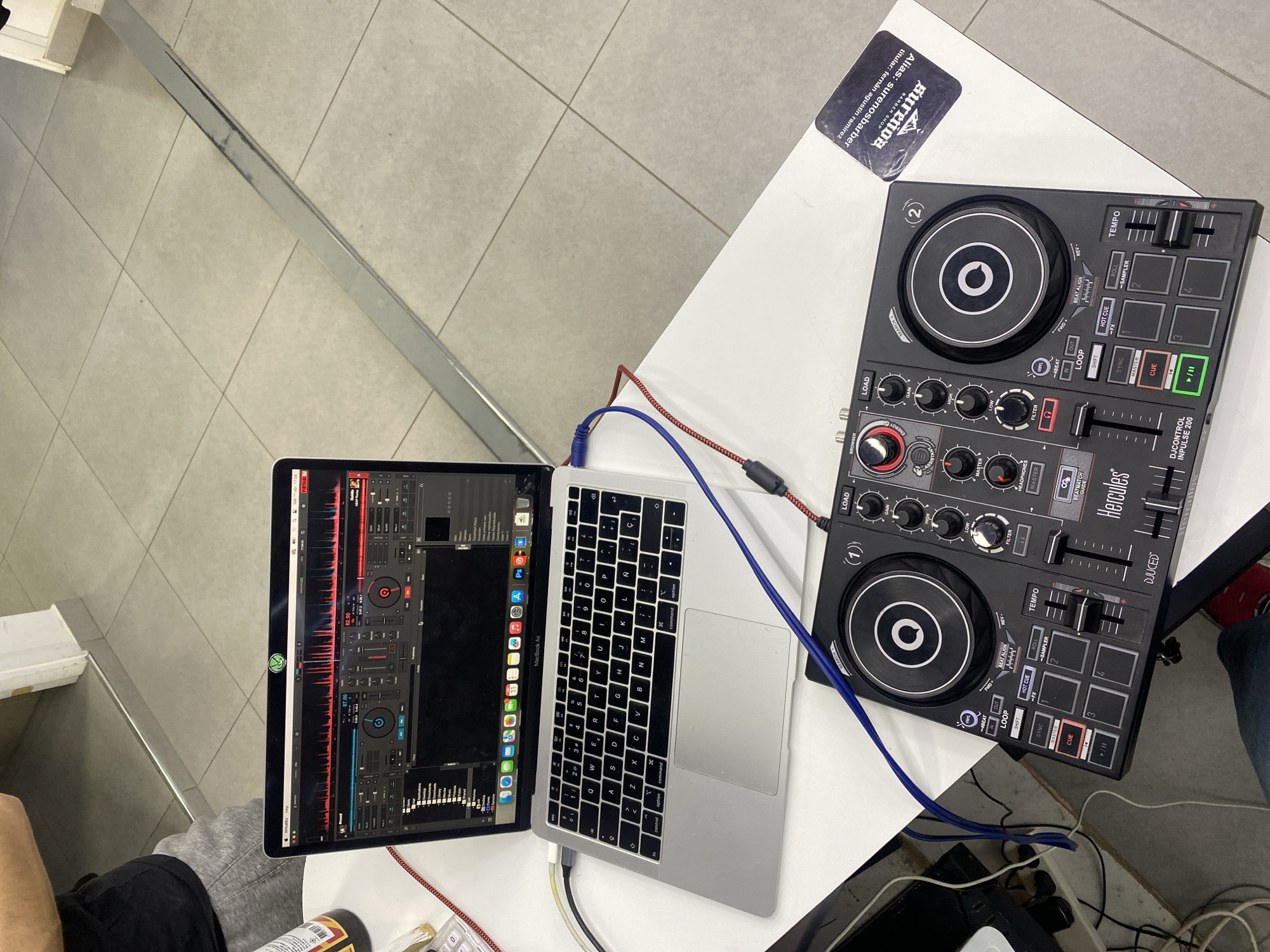 A digital music production setup is situated on a white table. A MacBook Air with DJ software displayed on the screen is connected via cables to a Hercules DJControl Inpulse 200 DJ controller. A business card is visible next to the controller with the name "Sureños Barber Shop," indicating the location is likely associated with a barber shop. The card lists Ushuaia, Argentina. A person wearing gray sweatpants is seated at the table and another person is visible wearing blue jeans with red shoes. Additional personal items, including a spray can and calculator, are present at the table. The table sits on a tile floor. A gray metal trim is also seen.