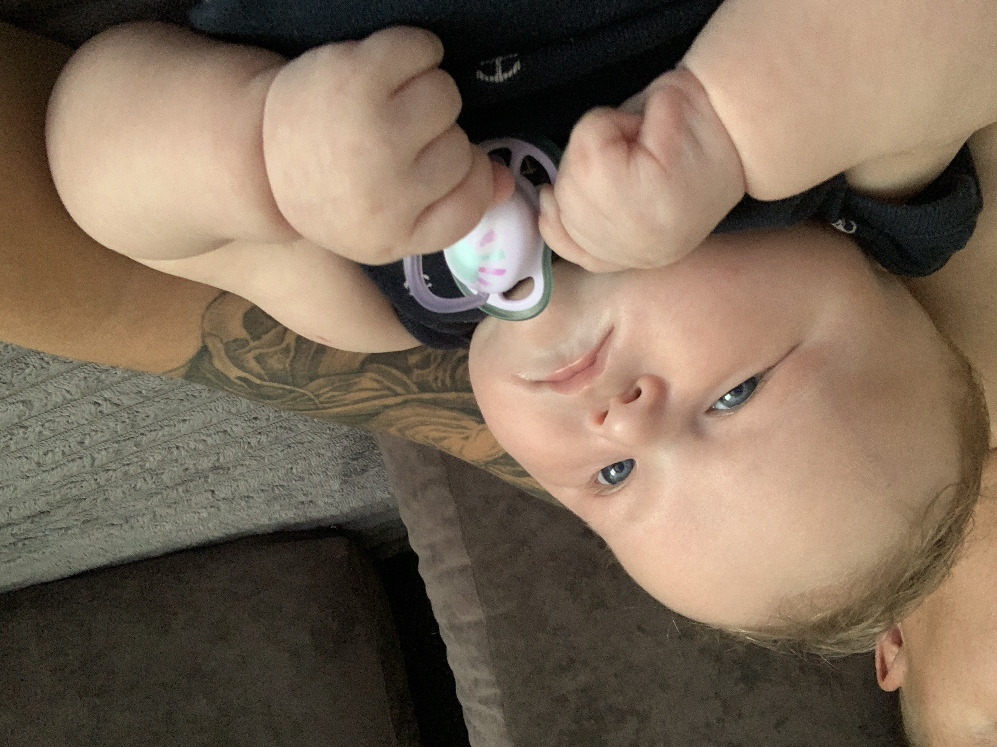 The image shows a close-up of a baby being held by a person. The baby, with blue eyes and fair skin, is wearing a dark blue onesie and holding a pacifier. The person holding the baby has a tattooed arm visible. They are likely on a couch or sofa, with a textured grey fabric visible in the background. The scene appears to be indoors, in a domestic setting. There is no information within the image that would allow for its specific location in Stavropol, Russia.