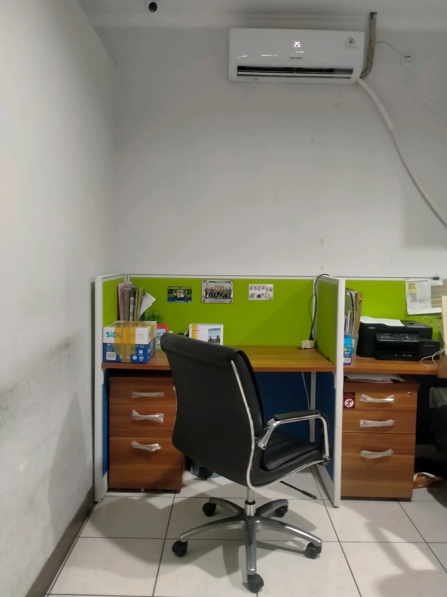 The image depicts a cubicle workstation in an office setting, likely in Ambon, Indonesia, given the context. The scene is viewed from a slightly elevated, frontal perspective. The workstation features a wooden desk with two sets of drawers on either side. On the left, a light brown drawer unit with three drawers is visible, with clear plastic coverings on the handles. On the right, a similar wooden drawer unit is present, also with three drawers and plastic-covered handles, along with a black Brother printer on top.
A black office chair with chrome accents and wheels is positioned in front of the desk. The cubicle walls are a light green fabric material. Pinned to the green wall on the left are several documents and a small framed photograph. On the desk to the left, there's a box of Sidu paper and some files. To the right of the chair, on the desk, a small calendar is visible.
Above the cubicle, a white split air conditioning unit is mounted on a white wall. The wall to the left of the cubicle has some discoloration or water stains. The floor is tiled with white square tiles. The overall impression is of a functional, albeit somewhat cluttered, office workspace.