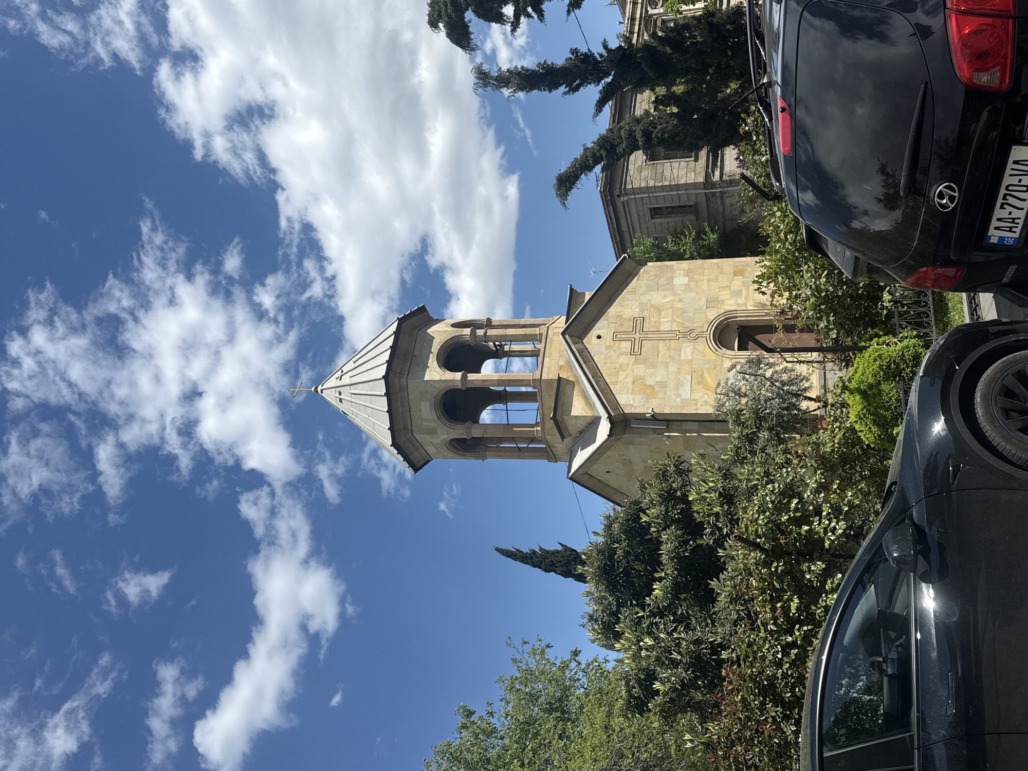 The image depicts the Anchiskhati Basilica, a church located in Tbilisi, Georgia, as it appears in the context of its urban setting.
The church’s architectural features, including its steeple and the cross on its facade, are visible against a backdrop of a bright blue sky with scattered clouds. Adjacent to the church are trees and some foliage. Two black cars are parked nearby. The composition of the image suggests a street-level perspective. The license plate of one of the vehicles is visible and shows the country of Georgia.