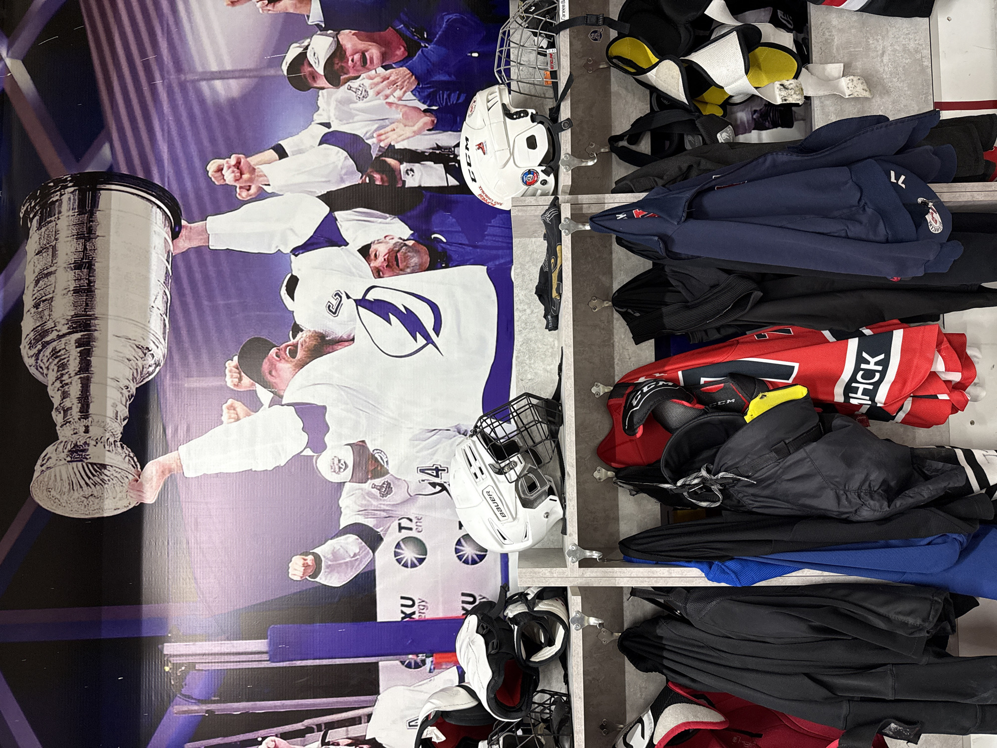 The image depicts a wall display, likely inside a sports facility, featuring a poster of the Tampa Bay Lightning hockey team celebrating with the Stanley Cup. The poster is oriented vertically, with the team members in white and blue jerseys hoisting the Cup. To the right of the poster are shelves containing hockey equipment, including helmets, jerseys, and pads. The jerseys appear to be various colors, including red, black, and blue. The specific location is not identified as Votkinsk, Russia.