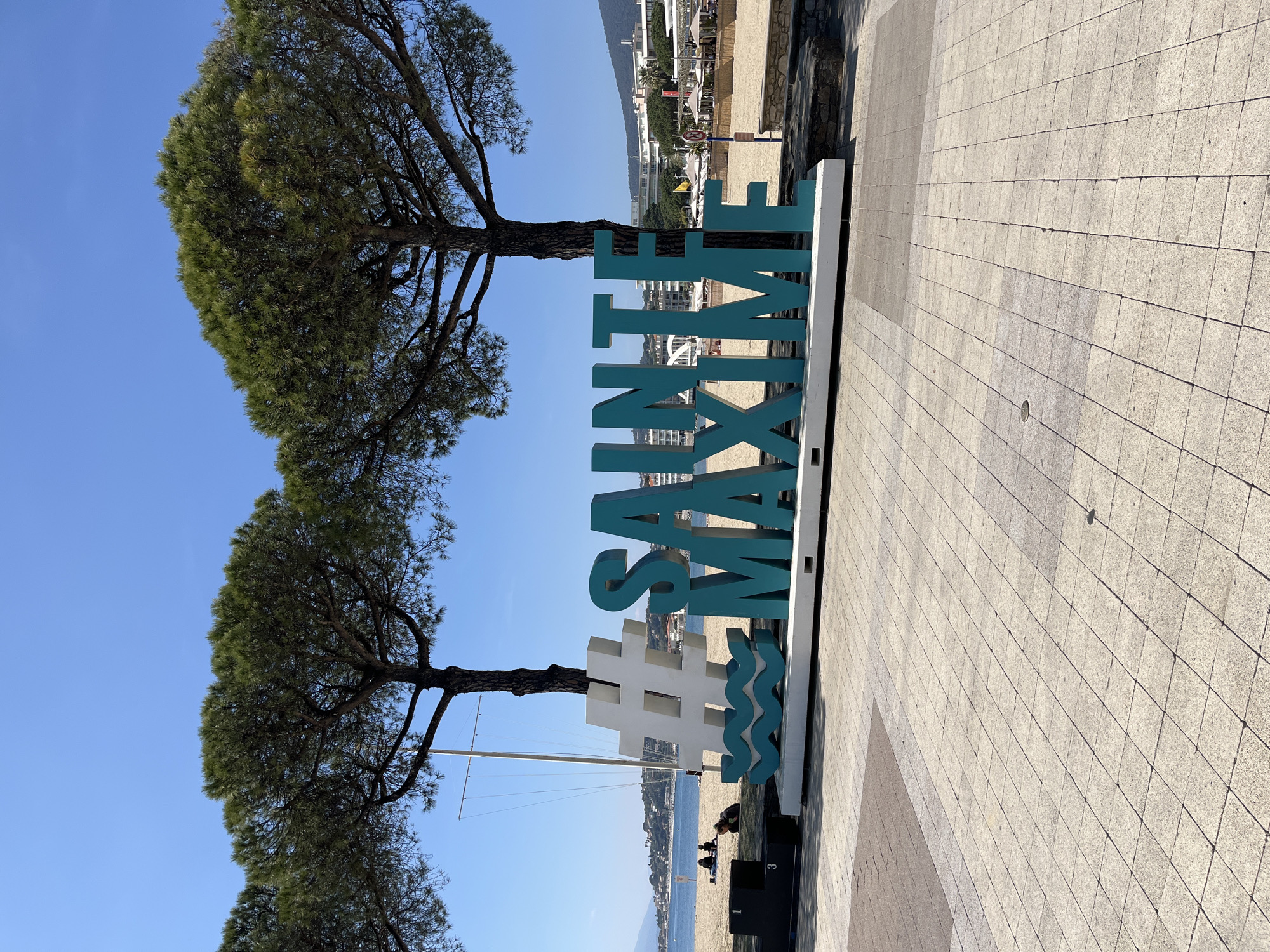 The image shows a sunny day at Sainte-Maxime, France. In the foreground stands a large, turquoise sign spelling "#SAINTE MAXIME" vertically, with wave designs underneath the hashtag. Two pine trees stand to the left of the sign. In the background is a beach with people, the Mediterranean Sea, and buildings. The ground is paved with square stones.