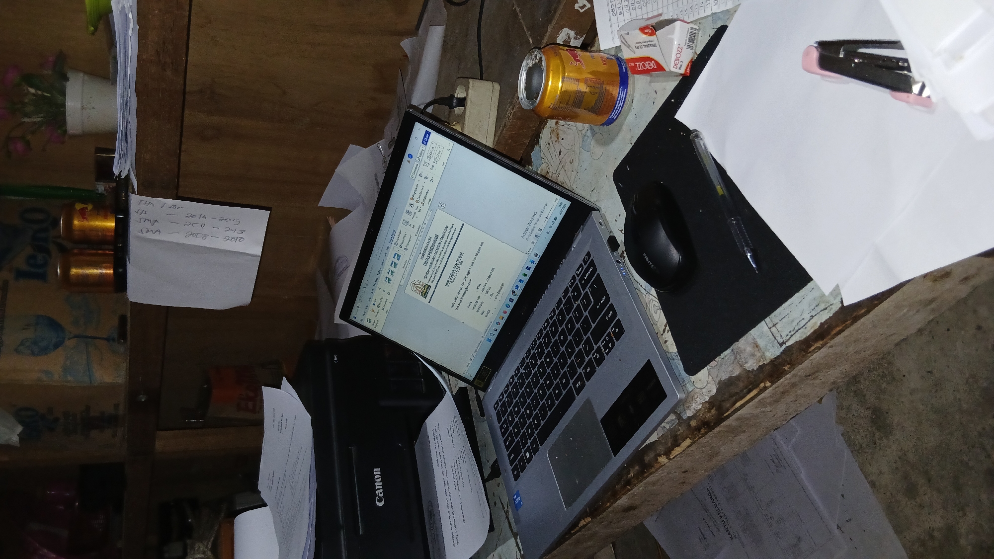 The image depicts a cluttered desk at a computer workstation. A silver laptop is open, displaying a document. To the left of the laptop, a black Canon printer is visible. A black computer mouse rests on a black mousepad. Papers are scattered across the desk and surrounding surfaces. A partially consumed can of Red Bull is situated to the right of the laptop, next to a small white box and some papers with handwritten text. In the background, shelves hold various items, including two golden cylindrical objects, a white potted plant with pink flowers, and cardboard boxes. The desk itself appears to be constructed from rough, weathered wood, and the floor beneath is concrete. The overall impression is of a work-in-progress or a recently used workspace. The location is identified as Keudejungkagajah, Indonesia.