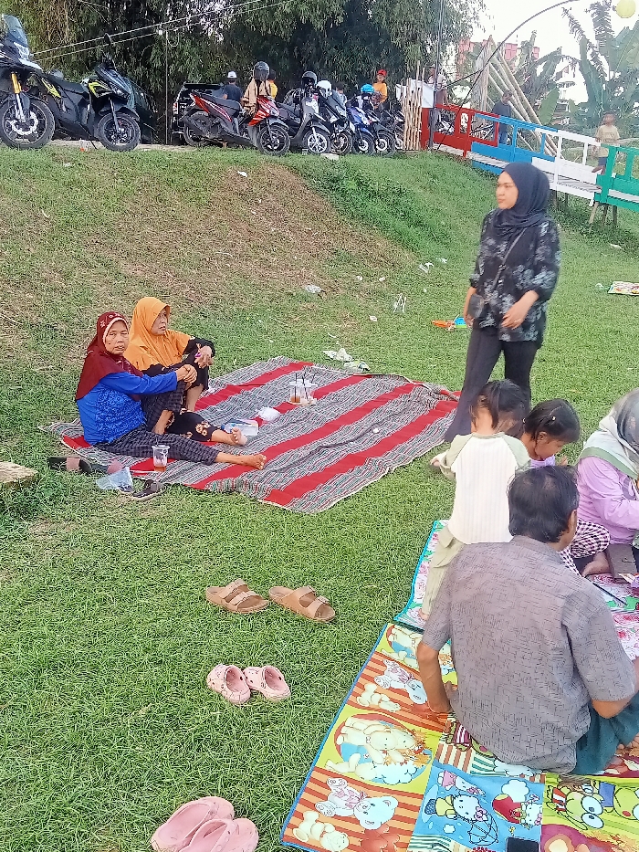 The image shows a group of people picnicking on a grassy hillside in Jakenan, Indonesia. The coordinates provided correspond to a location near the Jakenan River and a nearby park. There are three adults and three children present, all dressed in casual clothing. Two women are sitting on a red-and-white striped blanket, while the man is seated on a colorful, cartoon-themed mat, facing away from the camera. Three children are playing on the mat in front of the man. Two pairs of sandals are placed on the grass, and a third pair is nearby on the mat. Behind the picnic area, there is a row of parked motorcycles and a wooden bridge. Green grass covers the slope, and several trees are visible in the background.