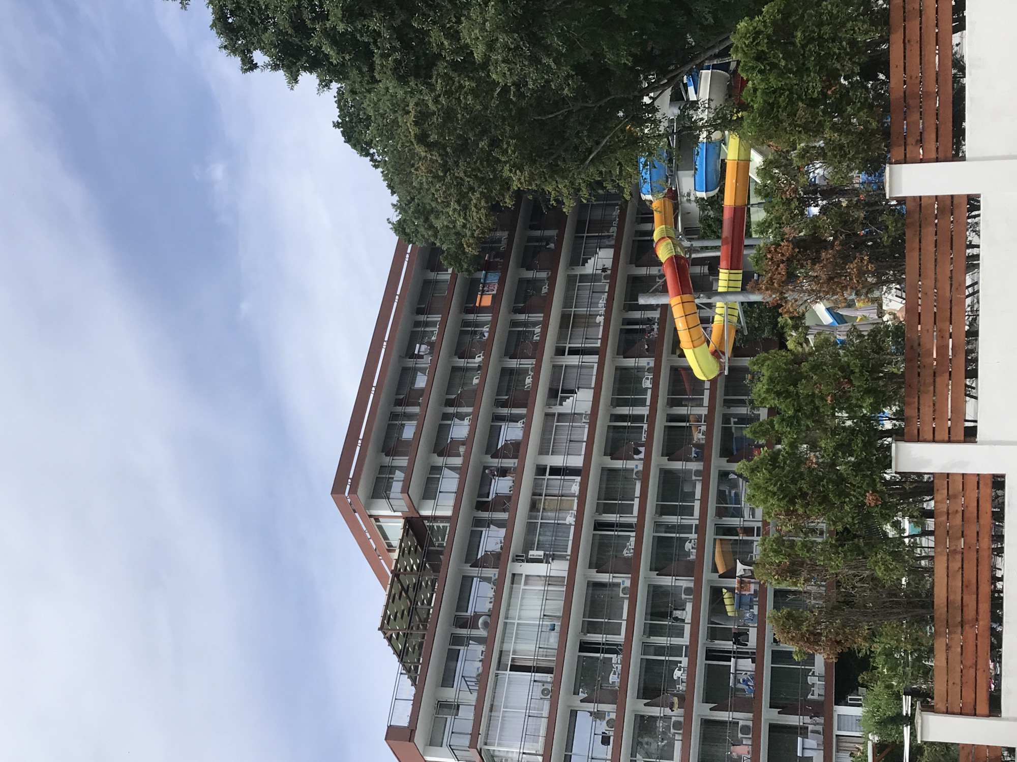 The image depicts a multi-story hotel with balconies and a water slide complex. The hotel is painted in white with reddish-brown accents around the balconies and the roofline. Several balconies are visible, furnished with chairs and tables, and some have towels or clothing hanging from them. Air conditioning units are mounted below some of the balconies. On the right side of the image, a large water slide with yellow, red, and blue sections is prominently featured, curving down from a higher level. In front of the hotel and water slides, there is a wooden fence with white posts. Lush green trees and bushes surround the area. The sky above is overcast with light clouds. The scene is likely located in a resort area, given the presence of the water slides and the hotel's architecture. The context suggests a leisure or vacation destination.