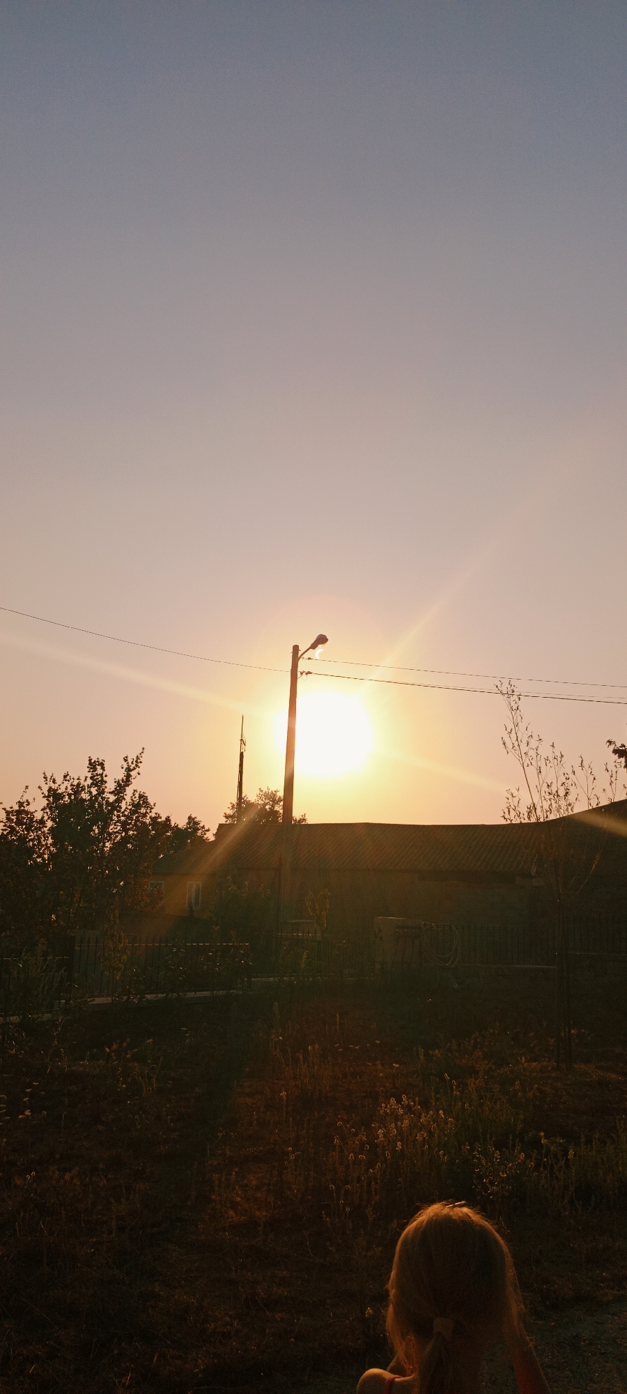 The image depicts a sunset with the sun prominently positioned near the center, casting a warm, orange glow. A silhouette of a young child, facing away from the camera, is visible in the lower right foreground. The child has blonde hair tied in pigtails. In the mid-ground, a house with a dark roof is visible, partially obscured by trees and vegetation. A wooden utility pole with electrical wires stretches across the scene. The overall environment appears to be a rural or suburban area, possibly on the outskirts of Adai, Spain, given the presence of houses and trees. The lighting suggests it is late afternoon or early evening.