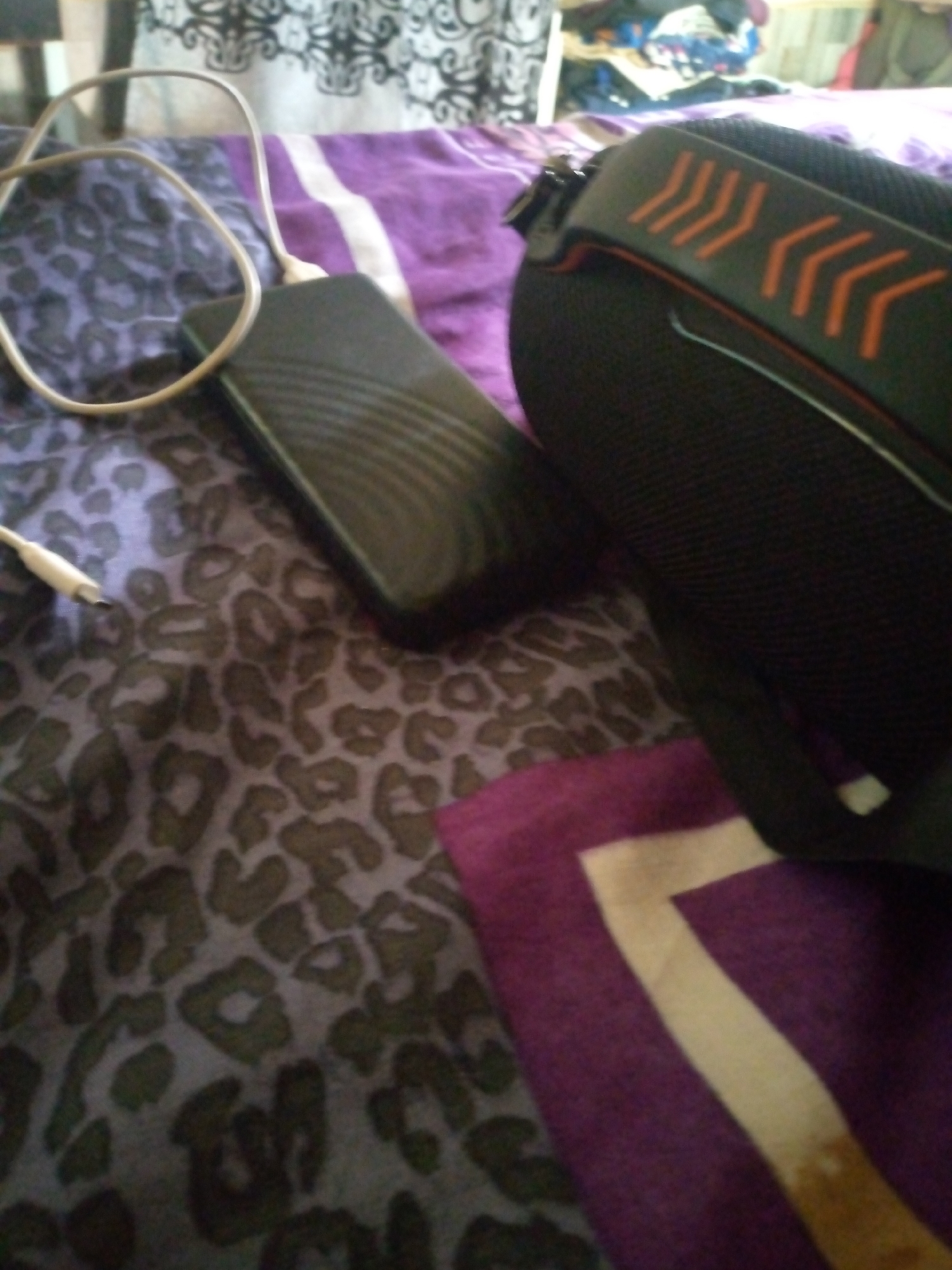 The image depicts a close-up, slightly blurry view of a bedside surface with a leopard-print fabric covering. On this fabric lies a black power bank connected to a white charging cable, with the cable forming a loop. Adjacent to the power bank is a black pair of headphones with orange chevron detailing on the headband. A purple fabric with a white stripe is also visible in the foreground. The background is out of focus but suggests an indoor setting with some stacked items. The image does not contain enough information to determine the specific location within Jalingo, Nigeria, or to describe any people, actions, or interactions.