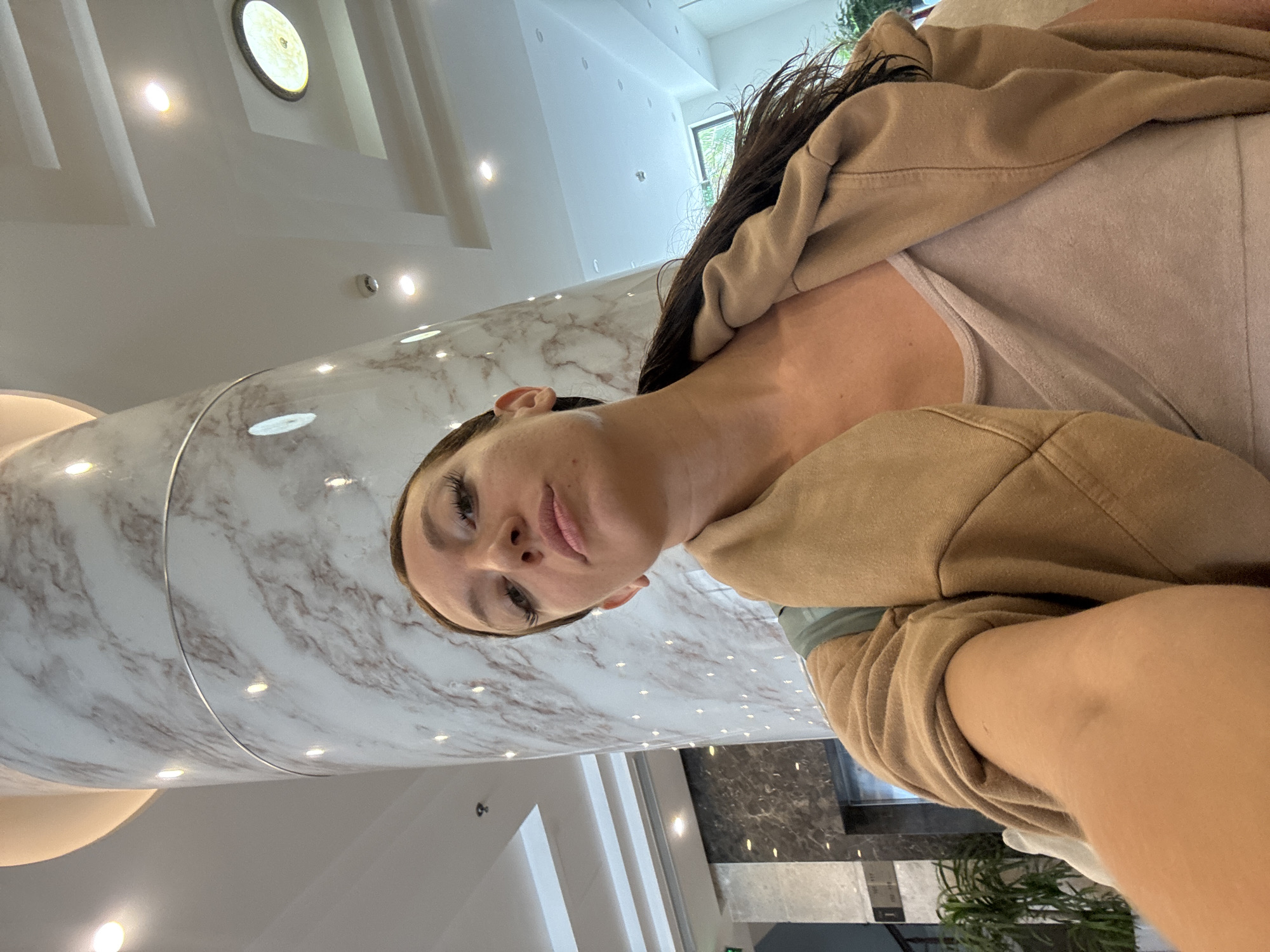 A selfie captures a woman from a low angle within a modern, upscale indoor setting, likely a hotel lobby or similar building in Đà Nẵng, Vietnam. The image is oriented vertically, with the woman's head towards the bottom left and her body extending diagonally across the frame.
The primary subject is a woman with long, dark hair, visible eyelashes, and full lips, looking directly into the camera with a neutral to slightly pensive expression, possibly a subtle pout. She is wearing a light-colored tank top or camisole under a loose-fitting, light brown or tan hooded jacket.
The background features striking architectural elements. A large, cylindrical column, clad in white marble with subtle grey and brown veining, dominates the left side of the frame. This column reflects numerous bright, circular recessed lights from the ceiling. The ceiling itself is white, adorned with several more recessed spotlights and a decorative, circular, backlit fixture in the upper left corner. Beyond the immediate foreground, white walls with additional recessed lighting and some architectural paneling are visible. In the distance, through what appears to be a large window or opening, bright daylight illuminates green foliage, suggesting it is daytime. Other parts of the background show darker, possibly grey or black, marble-like surfaces and indistinct structural elements.
The overall impression is one of a contemporary and well-lit interior, with polished surfaces and elegant design. The woman appears to be posing casually for a self-portrait. There is no visible text in the image.
