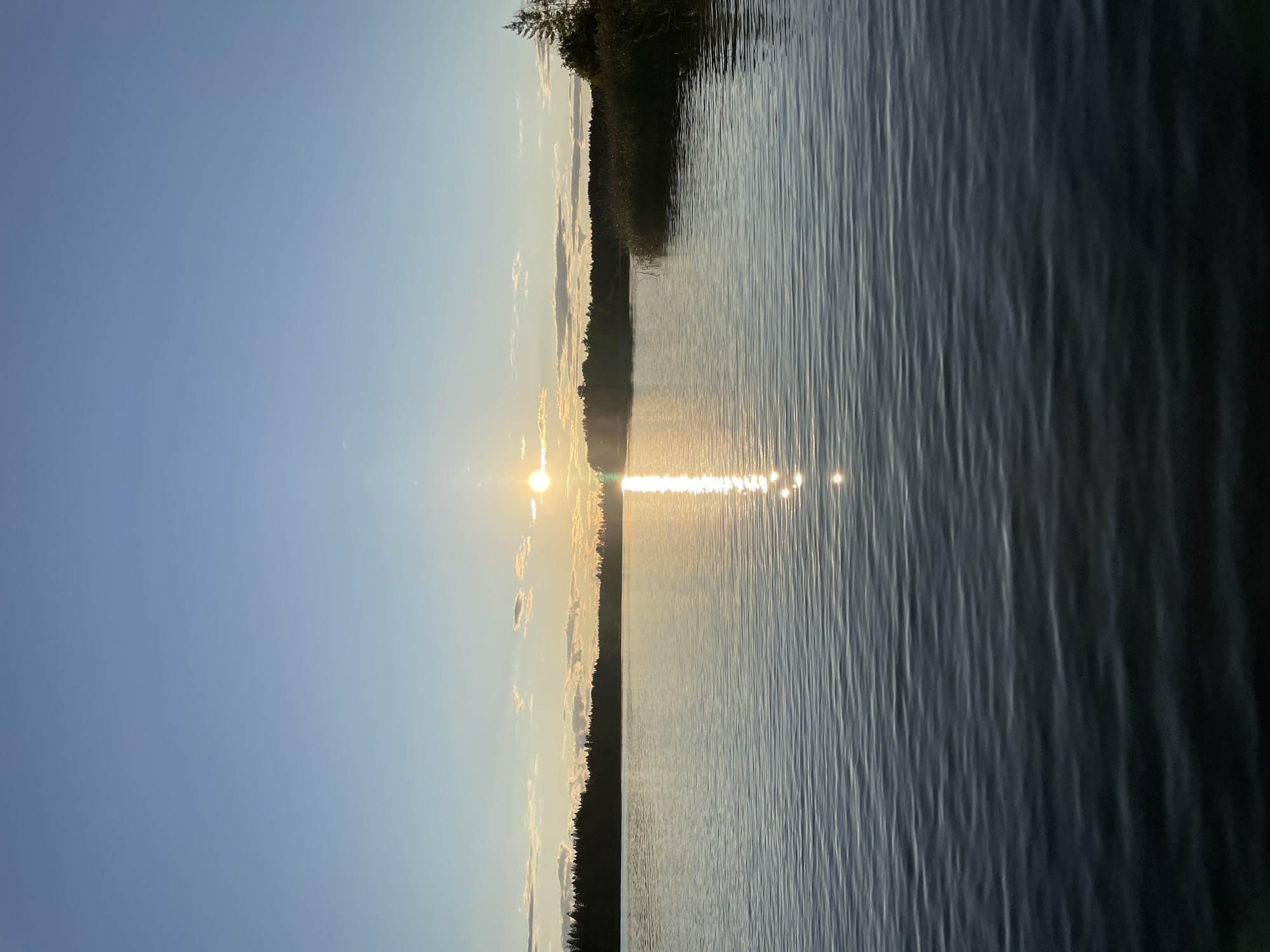 A vertical, wide shot captures a serene lake at sunset in Berozovo, Russia. The sun, a bright orb, is positioned in the upper center of the frame, casting a shimmering reflection on the water's surface. Wispy clouds dot the pale blue sky. A dense line of dark green trees forms a silhouetted horizon across the middle ground. The foreground is dominated by the rippling, dark blue water of the lake, reflecting the soft hues of the setting sun. No people or objects are visible in the scene. The overall atmosphere is one of tranquility and natural beauty.