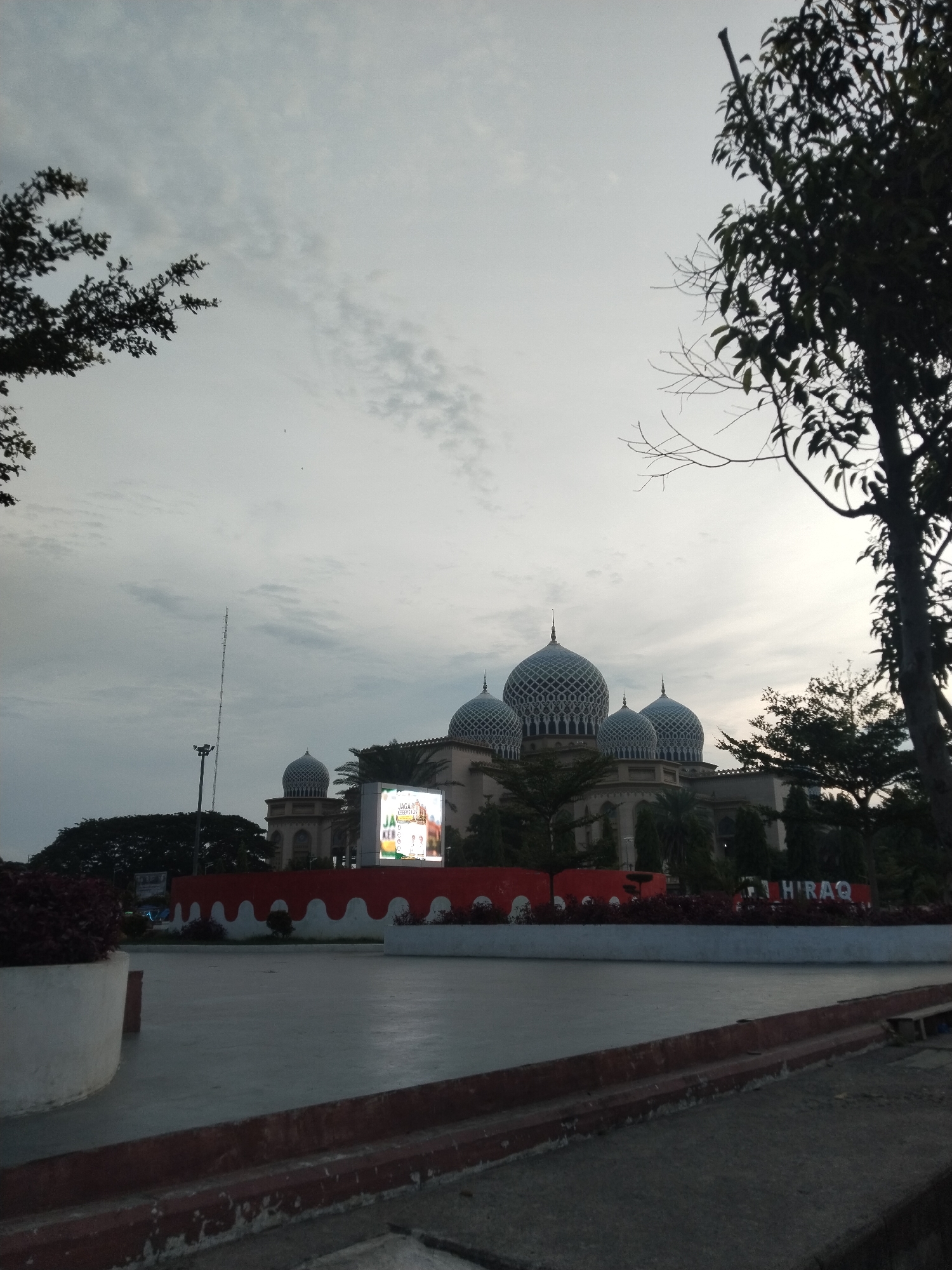 The image depicts the Baiturrahman Grand Mosque in the city of Lhokseumawe, Indonesia, under a cloudy sky. In the foreground, there is a paved plaza with a raised concrete border lined with red and white wavy patterns, and a large planter with dark red foliage on the left. A tall antenna tower is visible in the background to the left of the mosque. The mosque itself features multiple domes with intricate patterns, and a large LED screen displays an image with Indonesian text. Trees are present in the midground and foreground, framing the scene. The overall lighting suggests it is either dawn or dusk.
