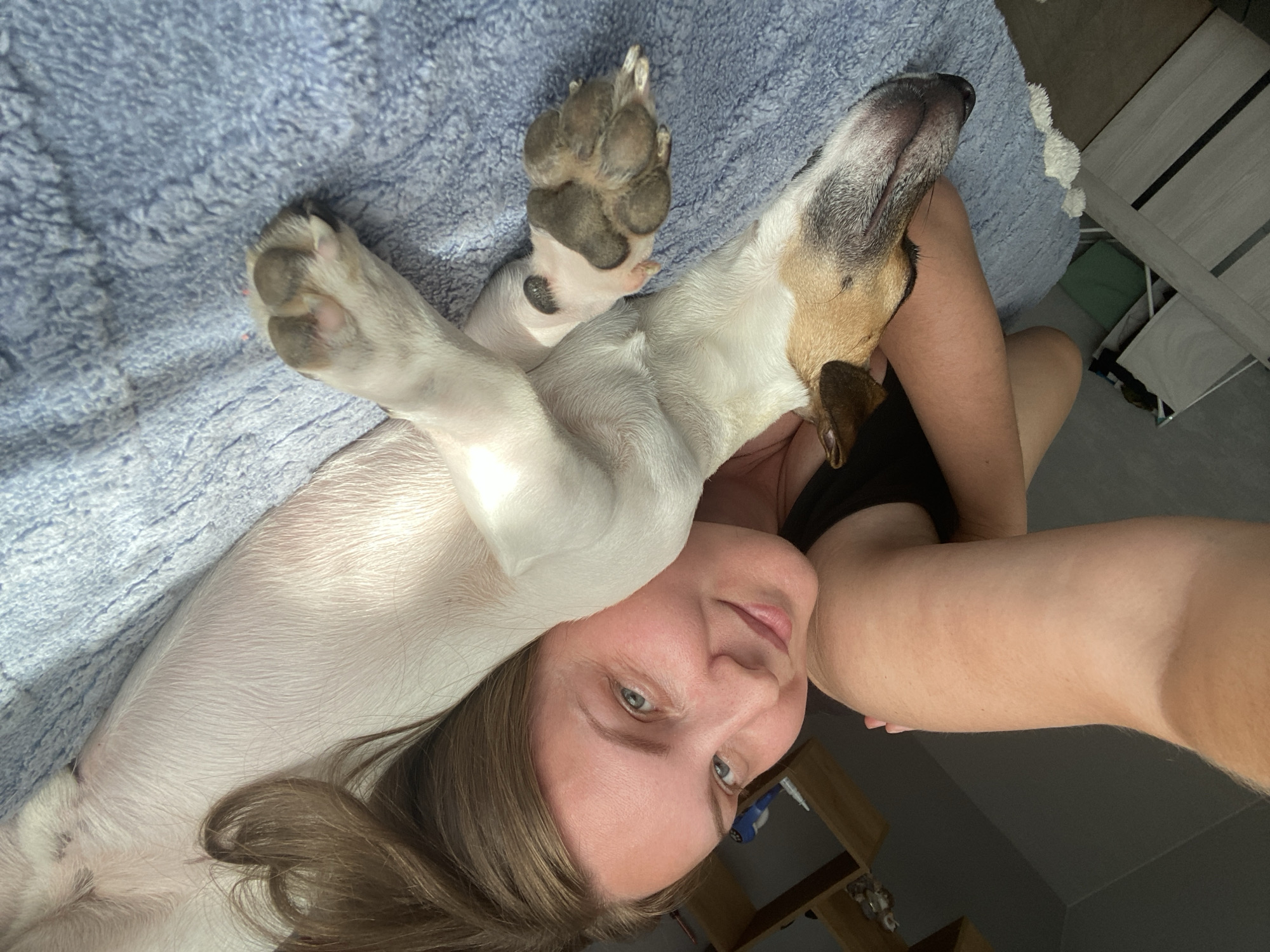 The image is a close-up, high-angle selfie taken indoors, depicting a woman and a dog lying together on a blue, textured blanket. The woman, with light brown hair and fair skin, is looking towards the camera with a slight smile. She is wearing a black tank top. Her left arm is extended, holding the camera.
The dog, a small breed with white and brown markings, is lying on its side, its head resting near the woman's face and its body extending across the frame. Its paws are visible, with one paw resting near the woman's arm. The background shows a wooden shelf with some objects, part of a white door, and a vertical blinds, suggesting an indoor domestic setting.
The image does not contain any visual information that would allow for the determination of the city of Noviy, Russia, or any specific urban location.