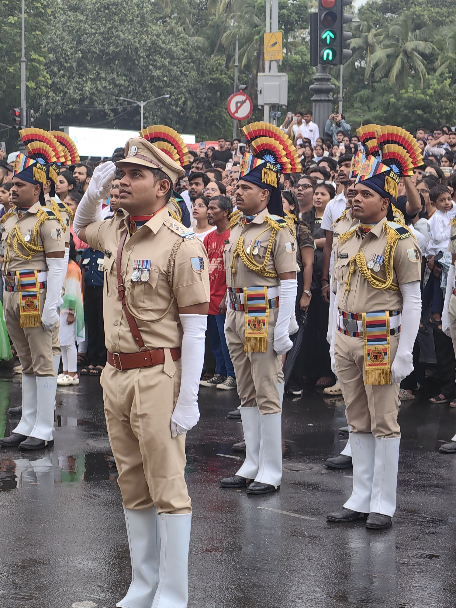 The image depicts a formal parade in Mumbai, India. Four men in police uniforms stand at attention, saluting. They wear tan uniforms with red collars, medals, and white gloves, and tall white boots. Their hats are adorned with colorful plumes. In the background, a crowd of spectators watches the procession. A traffic light with a green arrow is visible, suggesting an urban setting. The wet pavement indicates recent rain.