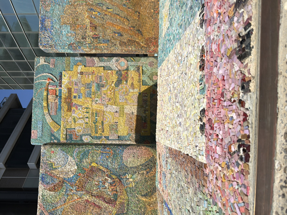 The image depicts a multi-paneled mosaic art installation, identified as Nachum Gutman's "Signon Tel Aviv" at Migdal Shalom (Shalom Meir Tower) in Tel Aviv, Israel. The installation features three prominent vertical mosaic panels composed of numerous small, irregularly cut tiles. The leftmost panel displays a complex design in blues, greens, and reds, depicting figures, a prominent crescent shape, and abstract forms. The central panel is predominantly yellow and gold, illustrating a stylized map or cityscape with geometric outlines, and an arc element at its top. The rightmost panel features a scene in earthy tones, with abstract or structural elements. In the foreground, a horizontal mosaic surface, likely a bench or ground element, is visible, segmented into distinct color blocks including pink, white, and blue-green tiles. Strong daylight illuminates the scene, casting defined shadows on the central vertical panel and across the foreground mosaic. Modern high-rise buildings with reflective glass and concrete exteriors are visible in the background under a clear sky. No individuals are present within the frame.