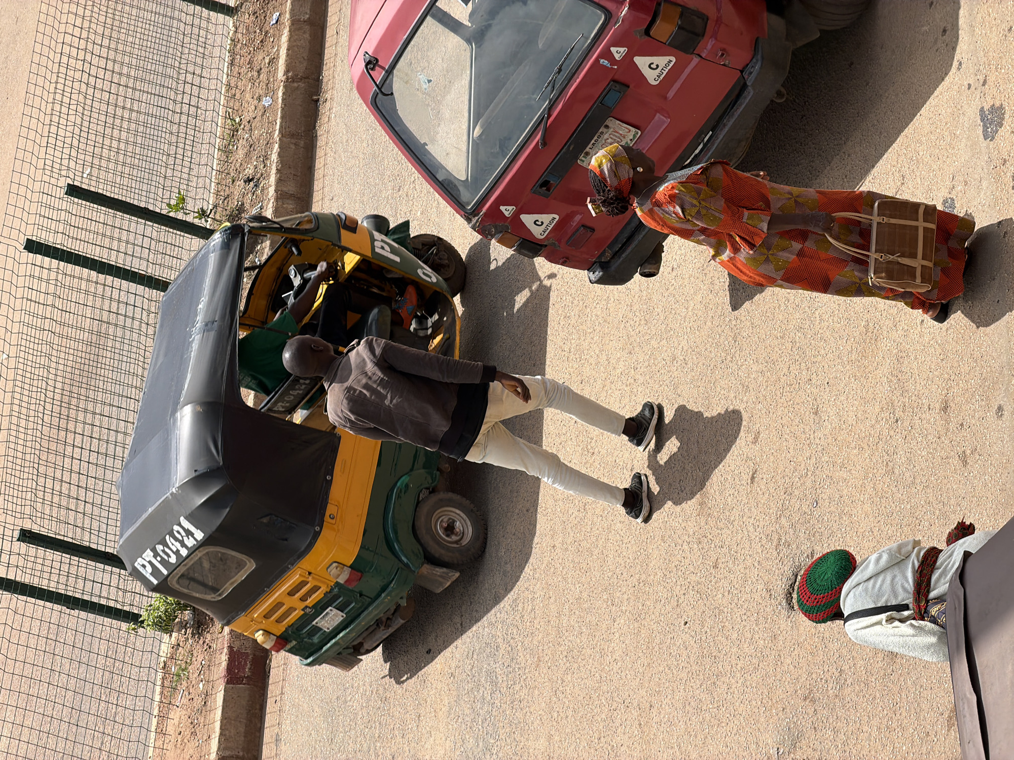 A person wearing a grey jacket and light-colored pants stands next to a yellow and green auto-rickshaw. Their legs are slightly bent, and they appear to be walking. To the right of the auto-rickshaw, a person in a vibrant orange and patterned dress carries a woven handbag. A red vehicle is parked behind them. In the background, a metal fence encloses a dusty area with sparse vegetation. The scene is illuminated by bright sunlight, casting sharp shadows, suggesting it is midday. The auto-rickshaw has "PT-0421" written on its side. The red vehicle has "LAGOS" visible on its license plate and a "CAUTION" sticker.
