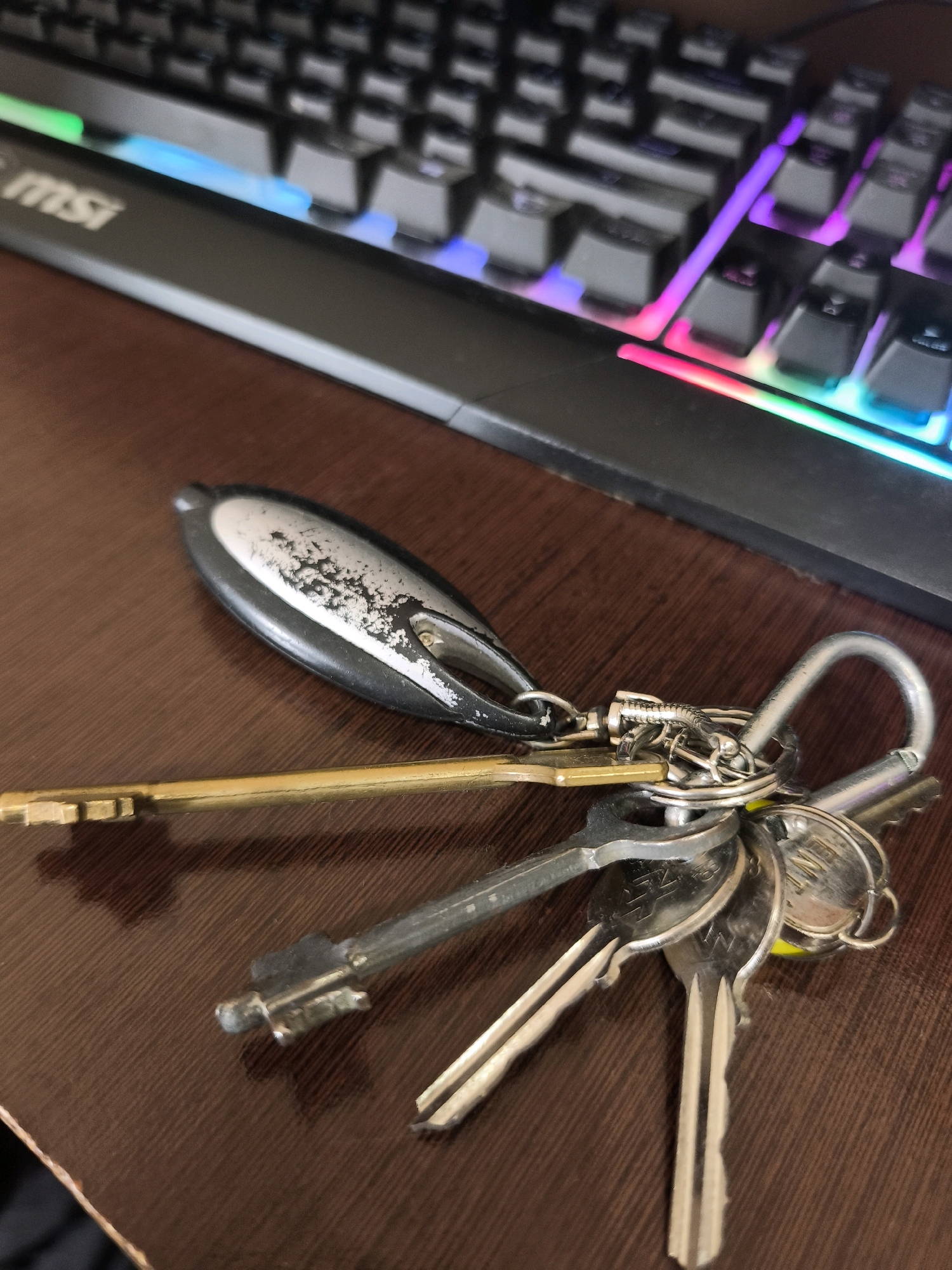 A close-up, high-angle shot captures a set of keys lying on a dark brown wooden surface, possibly a desk or table. The keys are attached to a silver carabiner clip, and among them is a gold-colored key. In the background, the lower portion of a black keyboard is visible, illuminated by vibrant, multi-colored RGB lighting that casts a glow onto the wooden surface and the keys. The keyboard has the "MSI" logo visible on its left side. The scene is devoid of any people or overt actions, presenting a still life of everyday objects. The image does not provide any information to suggest a location in the city of Karachayevsk, Russia.