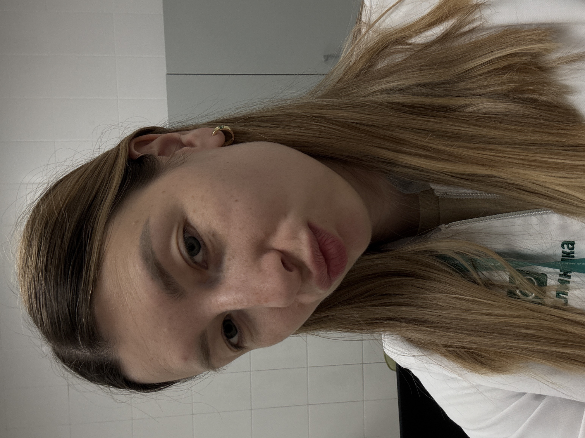 The image shows a rotated close-up portrait of a young woman, likely a selfie, captured indoors. Her head is tilted, and she gazes slightly downward and to her left, with a neutral expression and subtly pursed lips. Her long, light brown hair with blonde highlights frames her face and appears somewhat disheveled. Freckles are visible on her skin. She wears a small, gold-colored hoop earring with a green stone in her right ear.
She is dressed in a white uniform or lab coat, with a green lanyard or strap visible underneath her hair. The Cyrillic word "КЛИНИКА" (KLINIKA), meaning "Clinic," is clearly visible on this green strap and partially on the white uniform, strongly indicating a medical or health-related professional setting. The background consists of a white tiled wall and a reflective surface, possibly a mirror, reinforcing an indoor environment such as a clinic or hospital. The lighting is artificial and even. The presence of the word "КЛИНИКА" is consistent with the provided location context of Moscow, Russia.