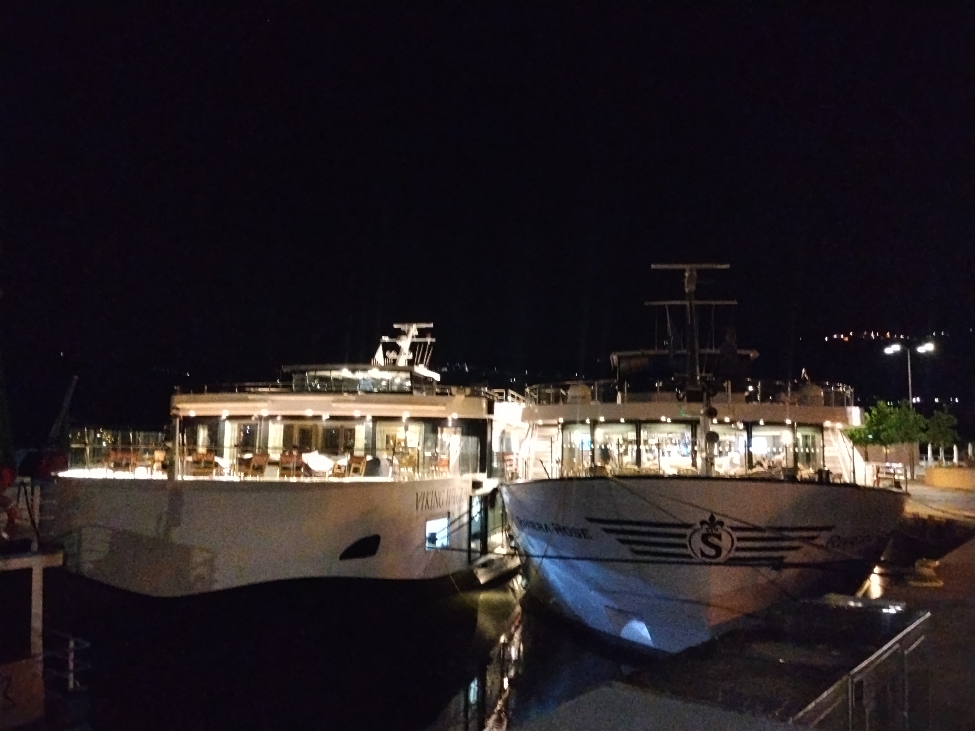 The image depicts two cruise ships docked at night in Peso da Régua, Portugal. The ship on the left is identified as "Viking Hachis", while the ship on the right is "Saborra Rose". Both ships are brightly lit and appear to be passenger vessels. Tables and chairs can be seen through the windows of both ships. The water is dark and reflects the lights of the ships. Streetlights are visible in the background on the right side of the frame.