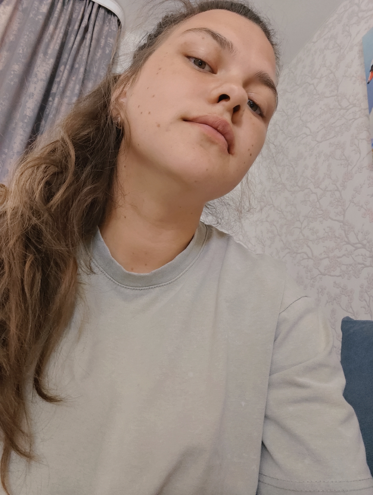 A woman with brown hair pulled partially back, wearing a light grey t-shirt, is the main subject of the image. She is positioned at an angle, looking down and slightly to the left. Her skin tone is light and she has some freckles. Behind her on the left is a grey curtain. The wall behind her on the right has a floral pattern. A blue cushion is visible in the lower right corner. The location is identified as Malyi Bukor, Russia.