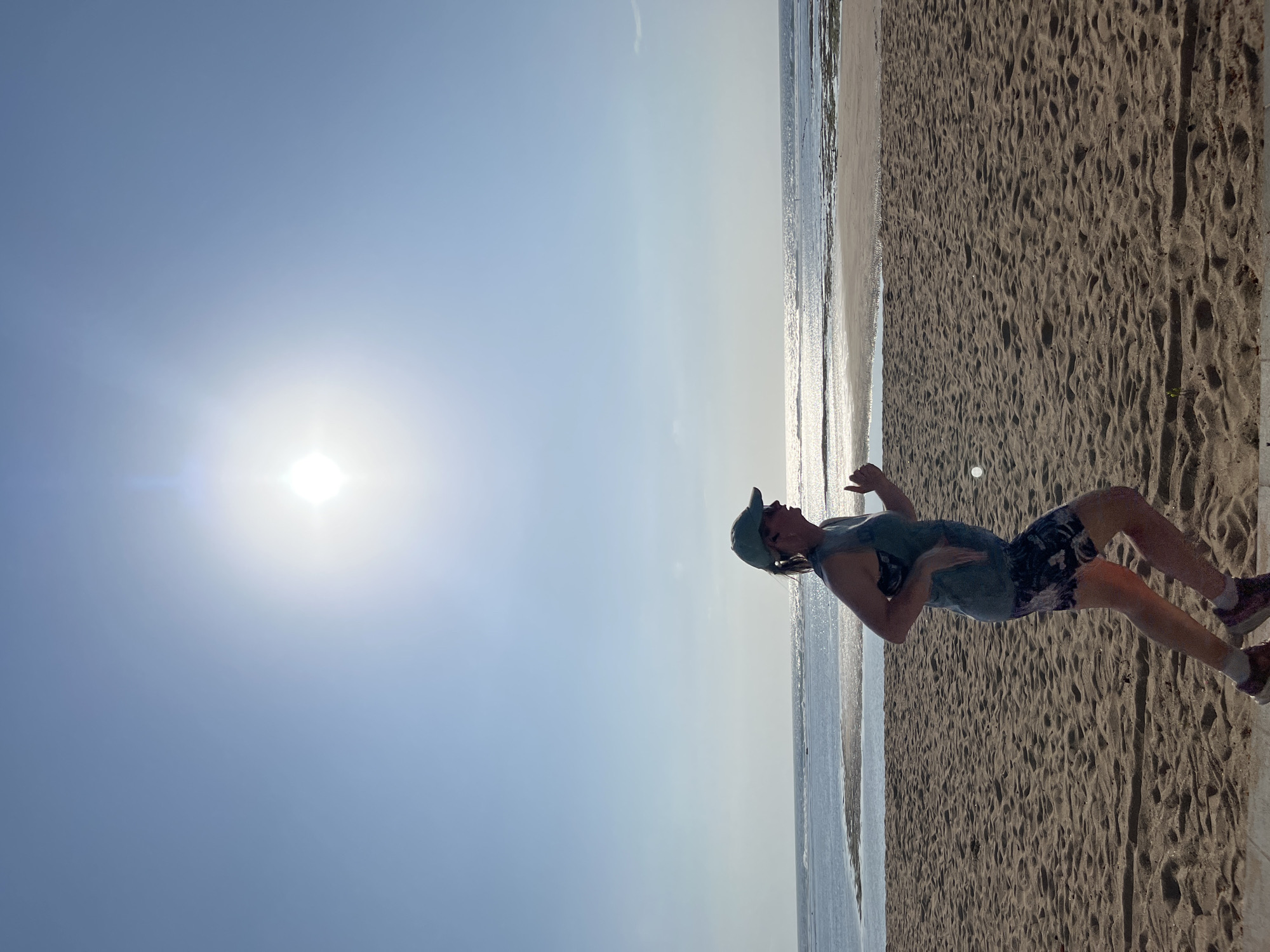 A full-body, side-profile shot captures a woman in athletic attire running on a sandy beach. The sun, a bright orb in the sky, is high and slightly off-center, casting a strong glare. The woman, wearing a teal tank top, patterned shorts, a teal baseball cap, and running shoes, appears to be in mid-stride, her right arm extended forward and her left arm bent at the elbow. Her gaze is directed forward. Behind her, the ocean stretches to the horizon, with gentle waves lapping at the shore. The sand in the foreground is textured with footprints and ripples, indicating recent activity. The sky is a clear, pale blue, with the sun being the dominant light source, creating lens flare. The scene appears to be set in Kuta Selatan, Indonesia, given the characteristic beach environment.
